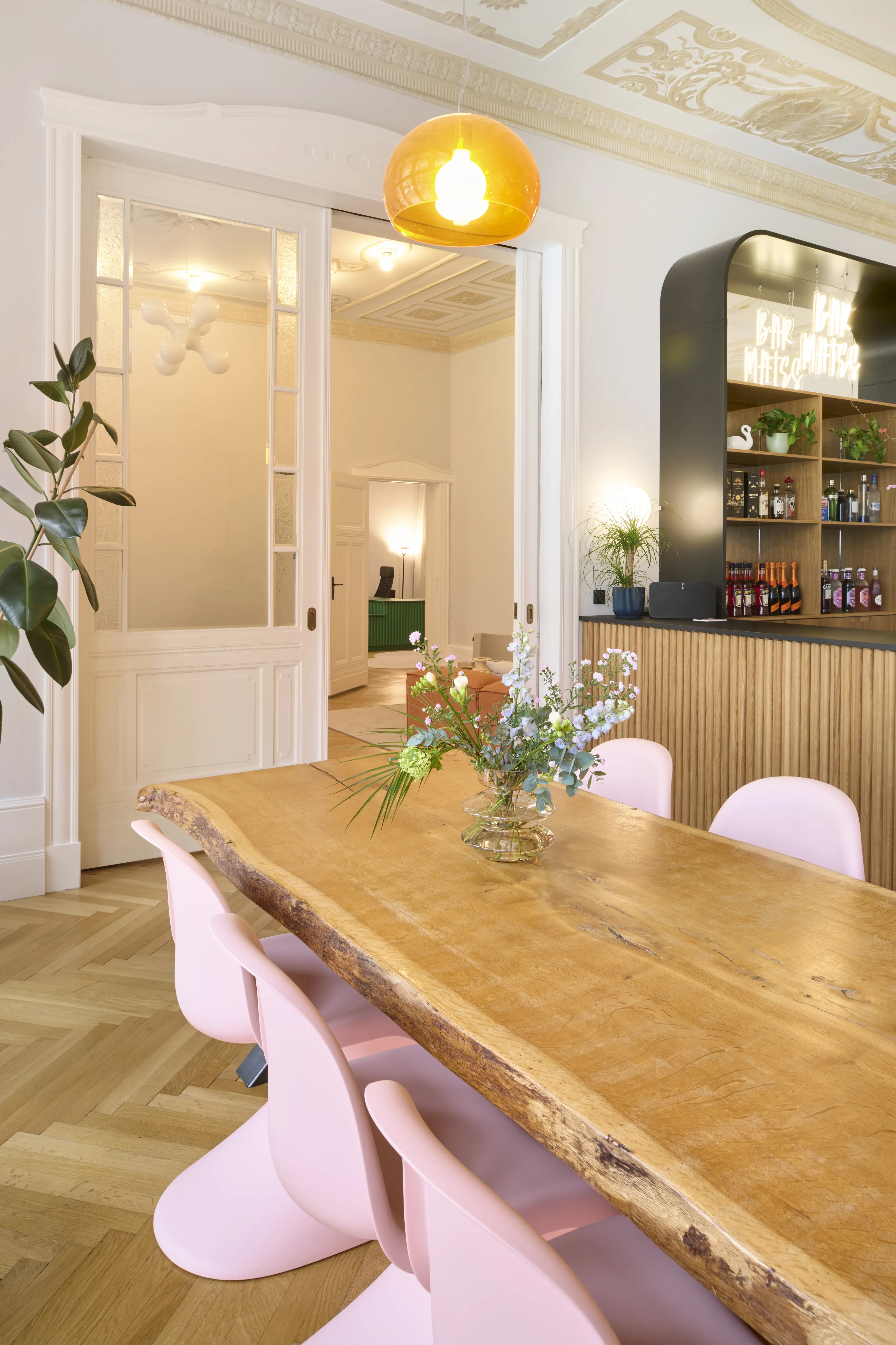 Dining room at INTERMATE BERLIN with a live-edge wooden table, pink Panton chairs, an amber glass pendant light, flower bouquet and the wooden bar with black arched shelf behind.