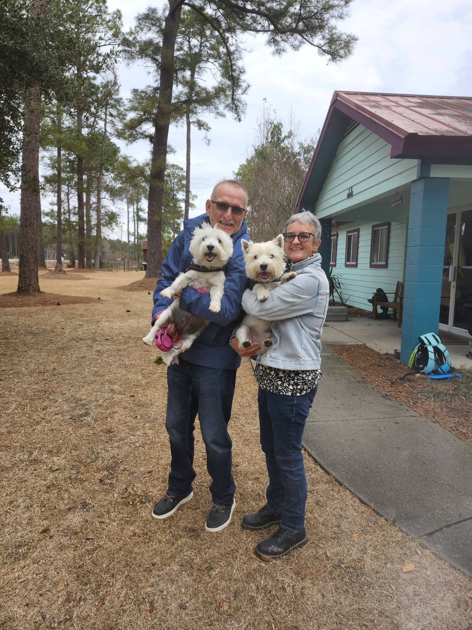 Two people stand outside a house, one holding a dog, with trees in the background and a cozy atmosphere.
