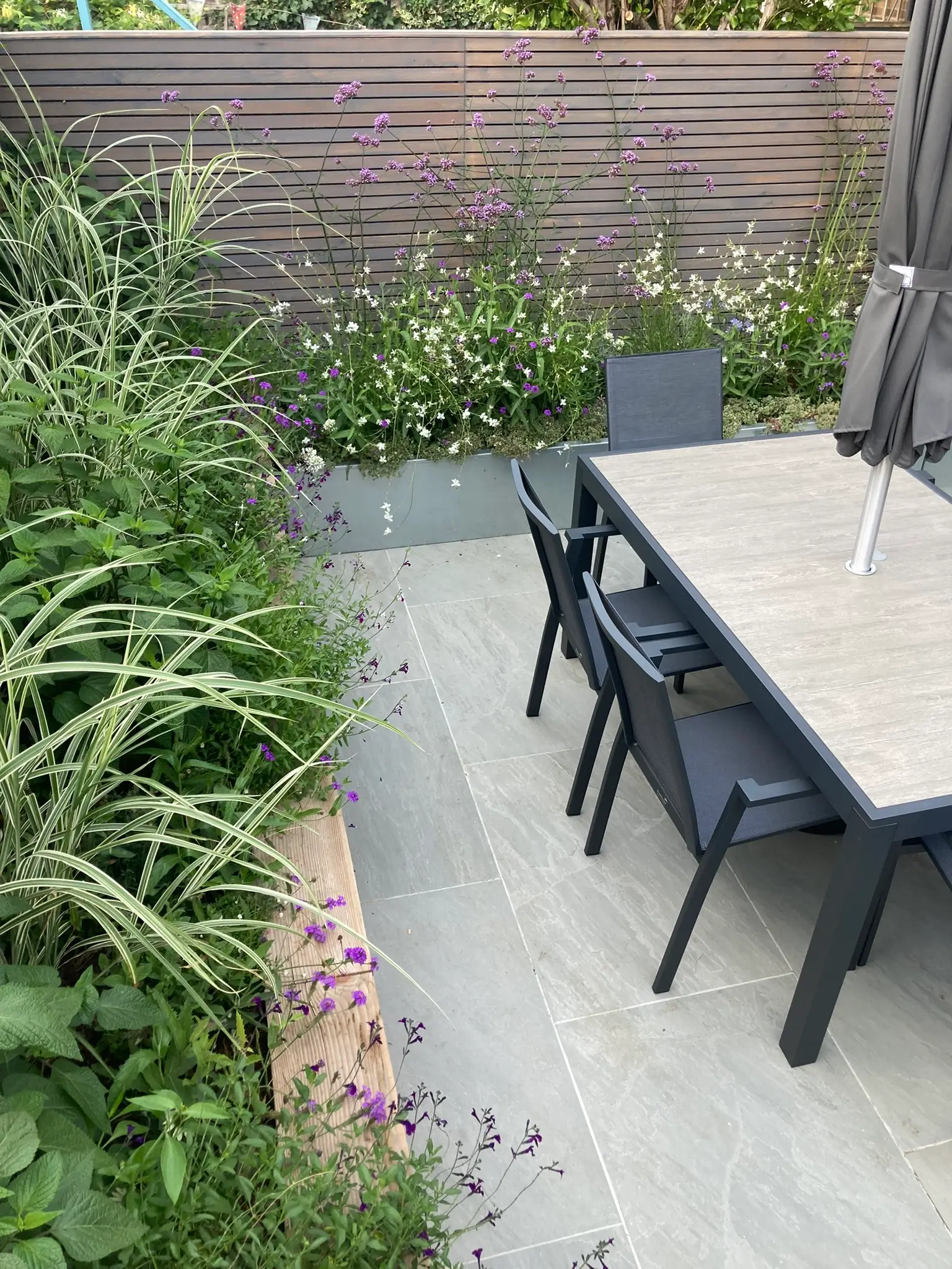 A garden patio featuring a table and chairs surrounded by lush greenery and a stone pathway.