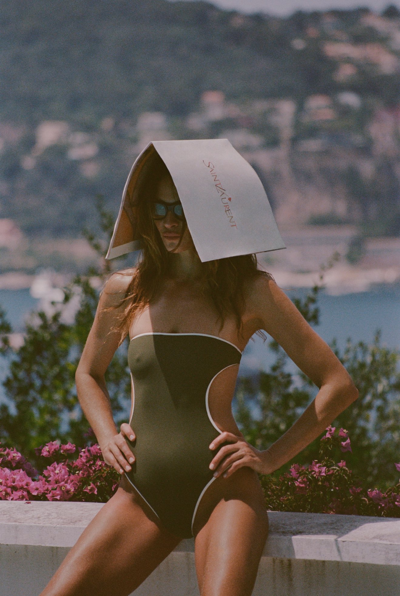 Model in an olive one-piece swimsuit poses outdoors by the sea for the Saint Laurent Summer 2025 campaign with a branded paper draped over her head..