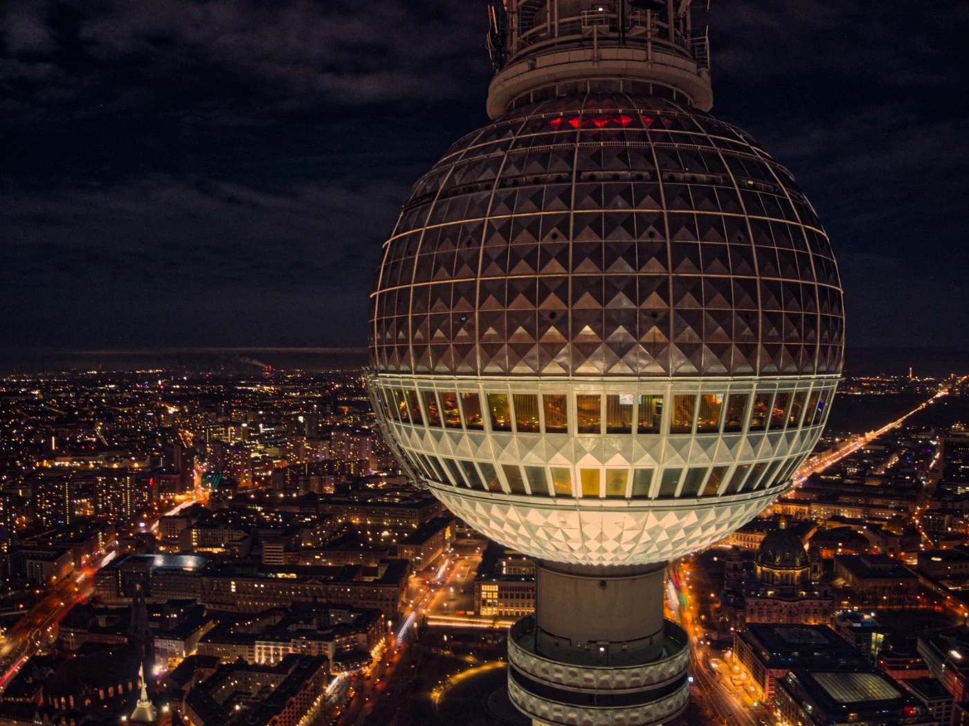 Nächtliche Drohnenaufnahme des Berliner Fernsehturms mit beleuchteter Kugel über dem Stadtzentrum, umgeben von weitläufigem Lichtermeer und Straßennetzen der Hauptstadt.