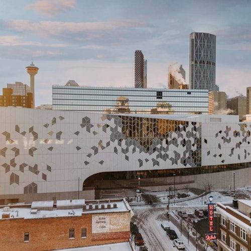 Cityscape with modern and older buildings, snowy streets, and a hotel sign. A unique building facade with geometric patterns is in the foreground.