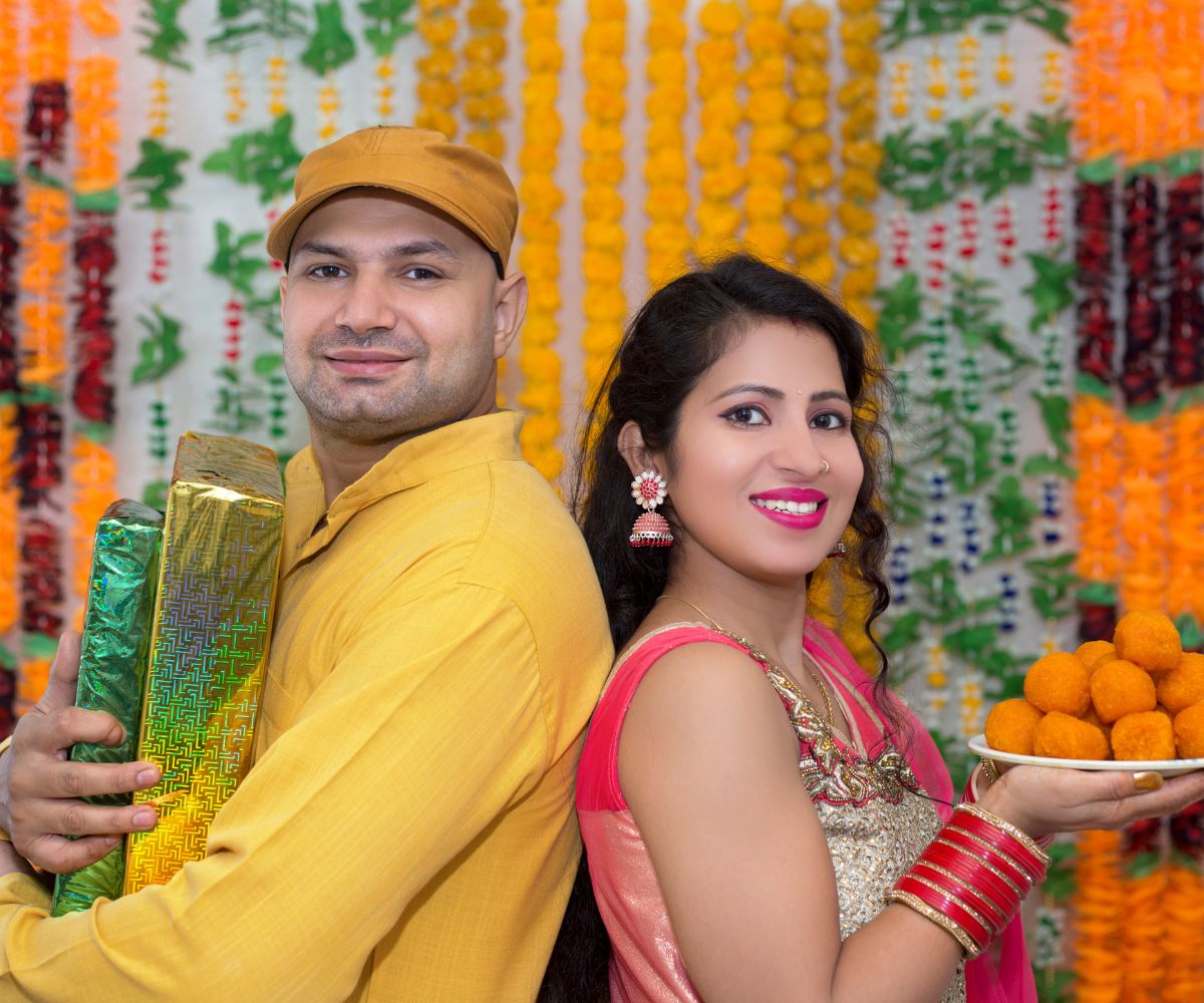 A cheerful duo in traditional attire celebrating Diwali at the office, surrounded by vibrant decorations. The man holds wrapped gifts, while the woman holds a plate of sweets, embodying the festive spirit, cultural significance, and joyous atmosphere of the Festival of Lights in a workplace setting.