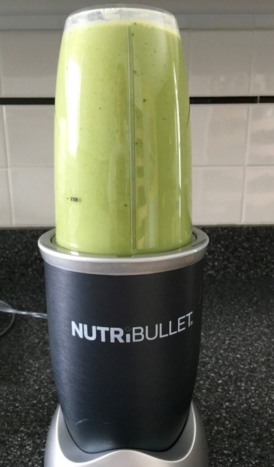 A NutriBullet blender sits on a kitchen countertop, blending a vibrant green smoothie, with white tile backsplash in the background.