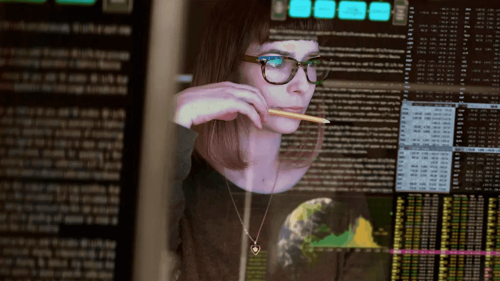 Woman in glasses studying data-filled screens in a darkened room, deep in focus.