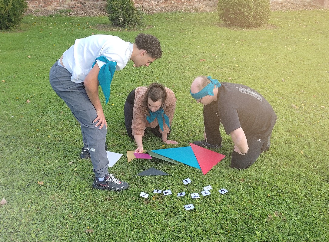 Trois personnes, un homme et une femme s'agenouillant pour résoudre une activité, tandis qu'un autre homme, debout, regarde attentivement. Ils sont en train de manipuler des pièces colorées (triangles et cartes) sur de l'herbe.