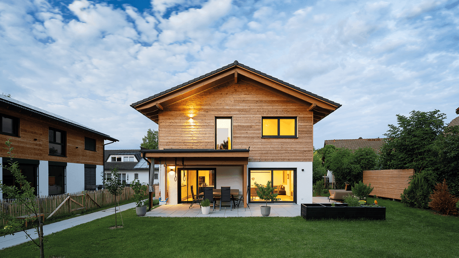 Modernes Holzhaus mit großen Fenstern und Gartenmöbeln auf der Terrasse bei Dämmerung.