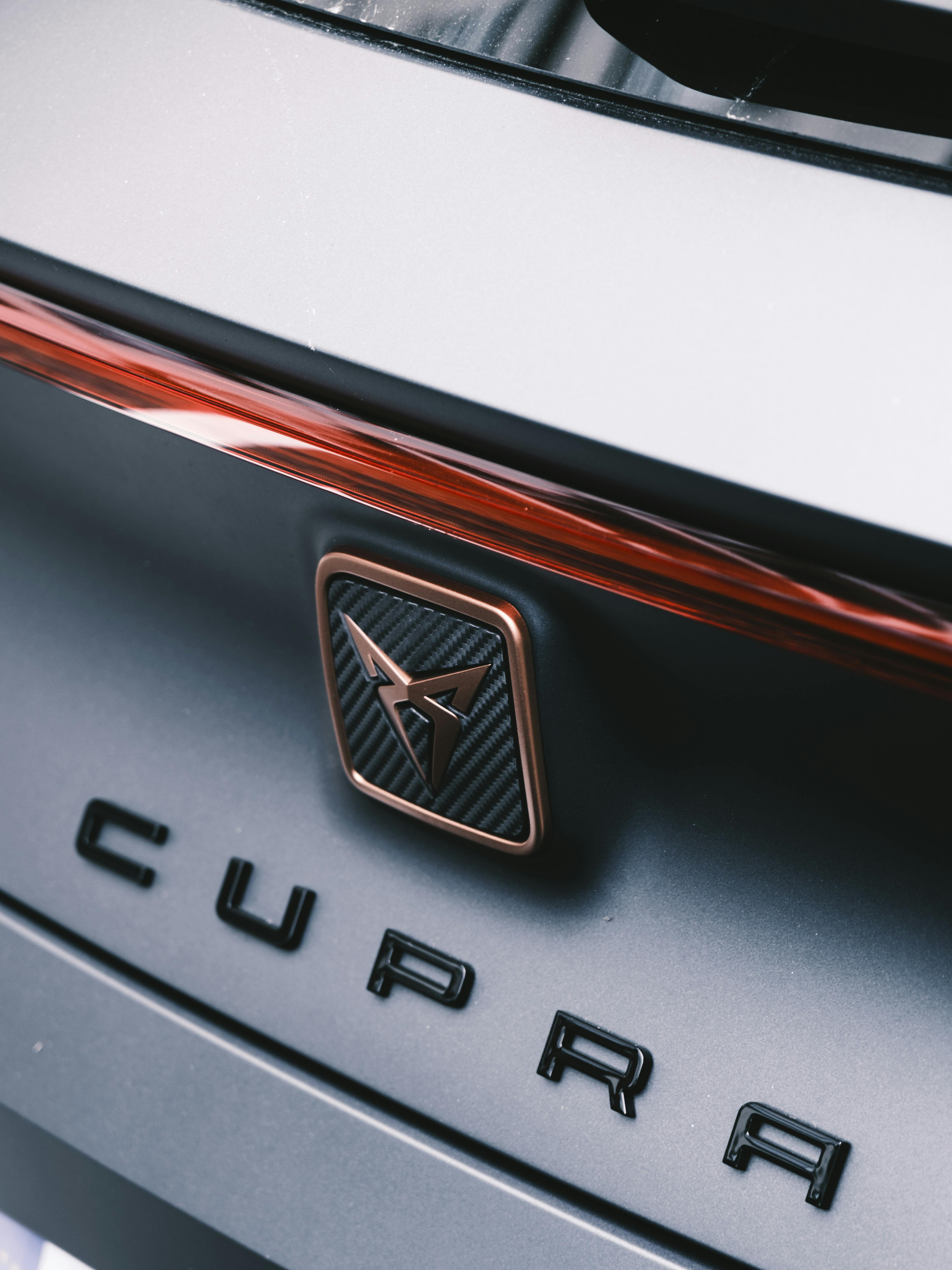 Photographie macro du logo Cupra cuivré posé sur un capot sombre, avec un flou artistique en arrière-plan qui accentue la profondeur et le relief du badge. Une mise en valeur élégante et minimaliste de l’identité visuelle distinctive de Cupra, symbole de performance et de design avant-gardiste.