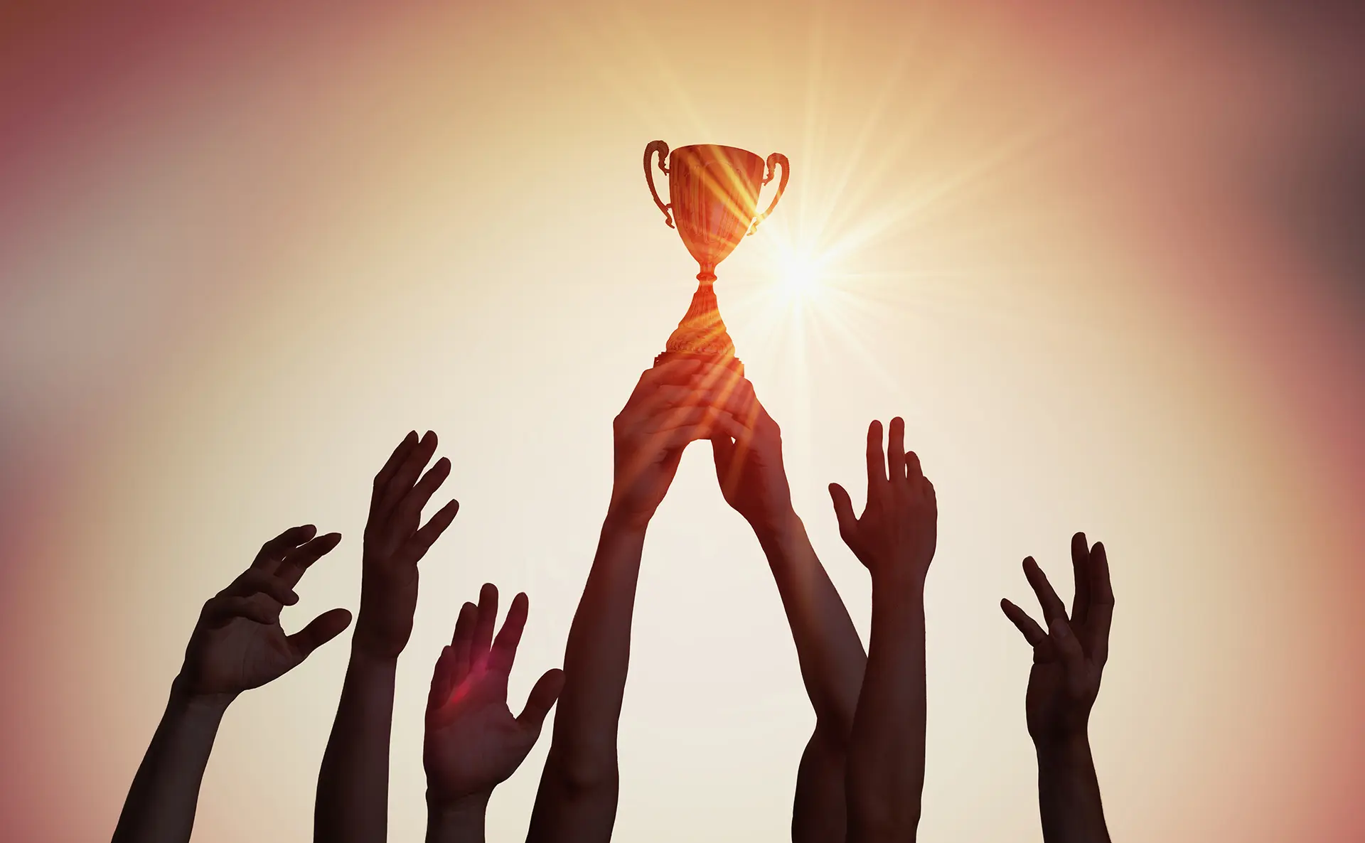 Multiple hands lifting a golden trophy against a bright sunset, celebrating a team achievement