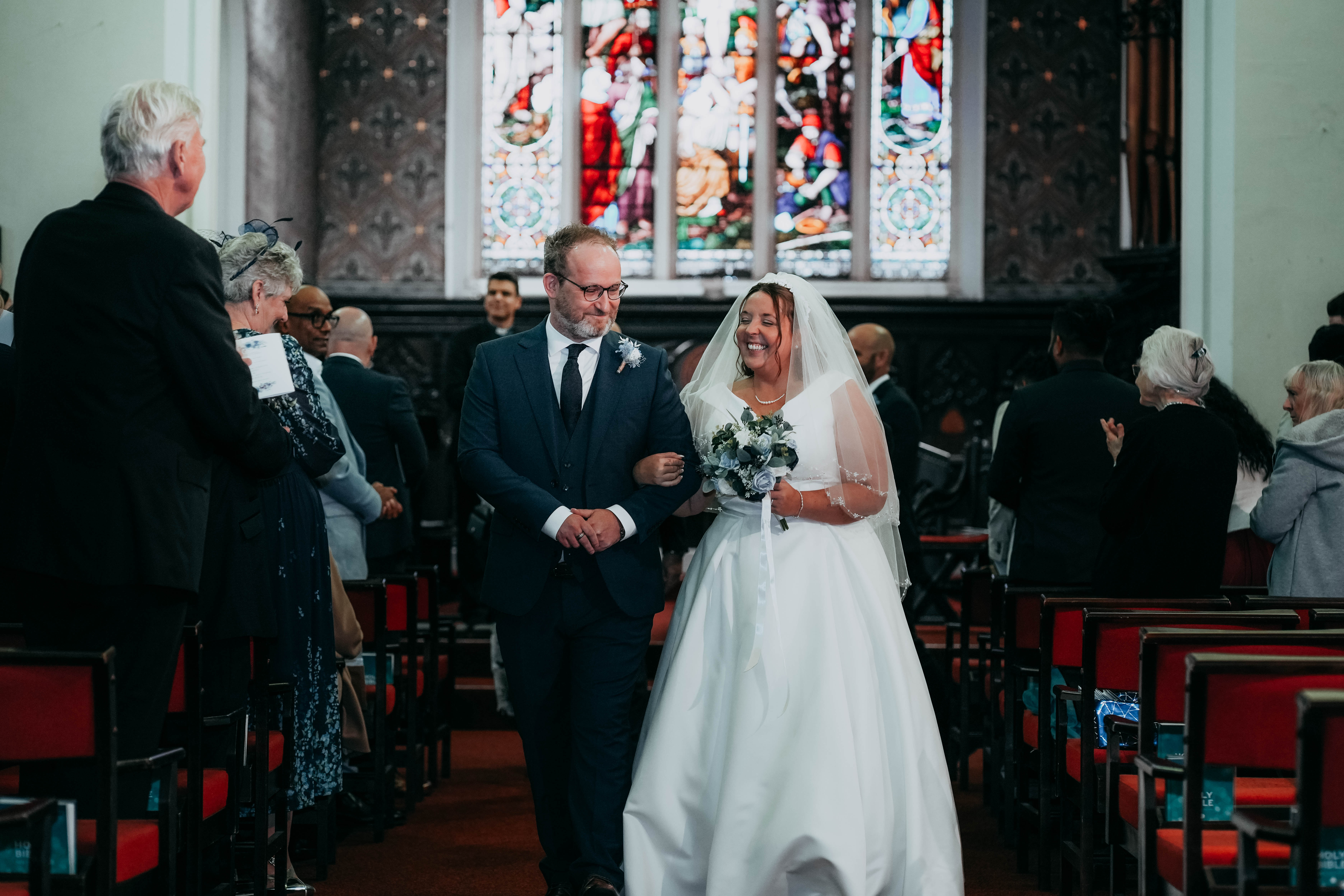 The newly married bride and groom walk arm-in-arm back up the church aisle, smiling at each other as guests look on. A large stained-glass window is visible behind them.