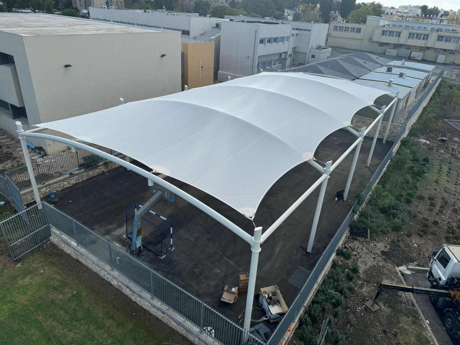 Large beige curved tent roof on metal frame over paved outdoor area next to buildings.