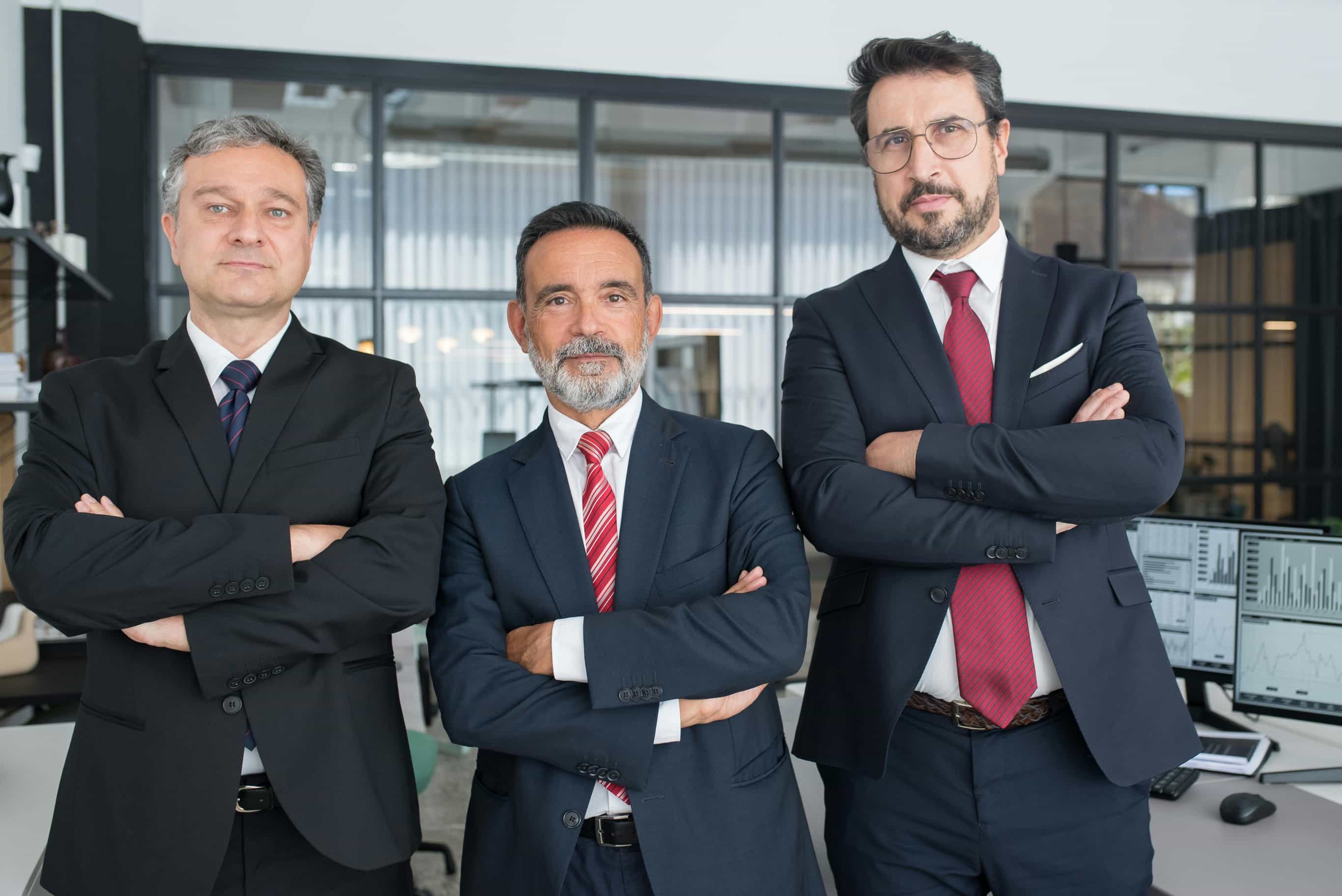 A group of three professional men in dark suits with ties (one red, one dark) stand side-by-side with their arms crossed, looking confidently at the camera in a modern office environment.