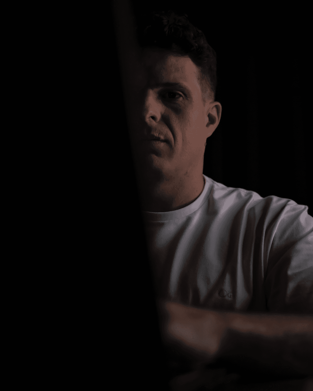 Man sitting in a dimly lit room, wearing a beige shirt and looking contemplative.
