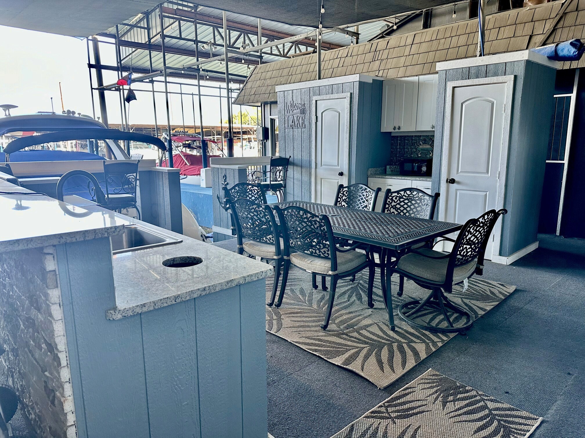 Outdoor dining area under a covered patio features a metal table with matching chairs on decorative rugs, adjacent to a kitchenette with a stone countertop, overlooking a marina with docked boats in the background.