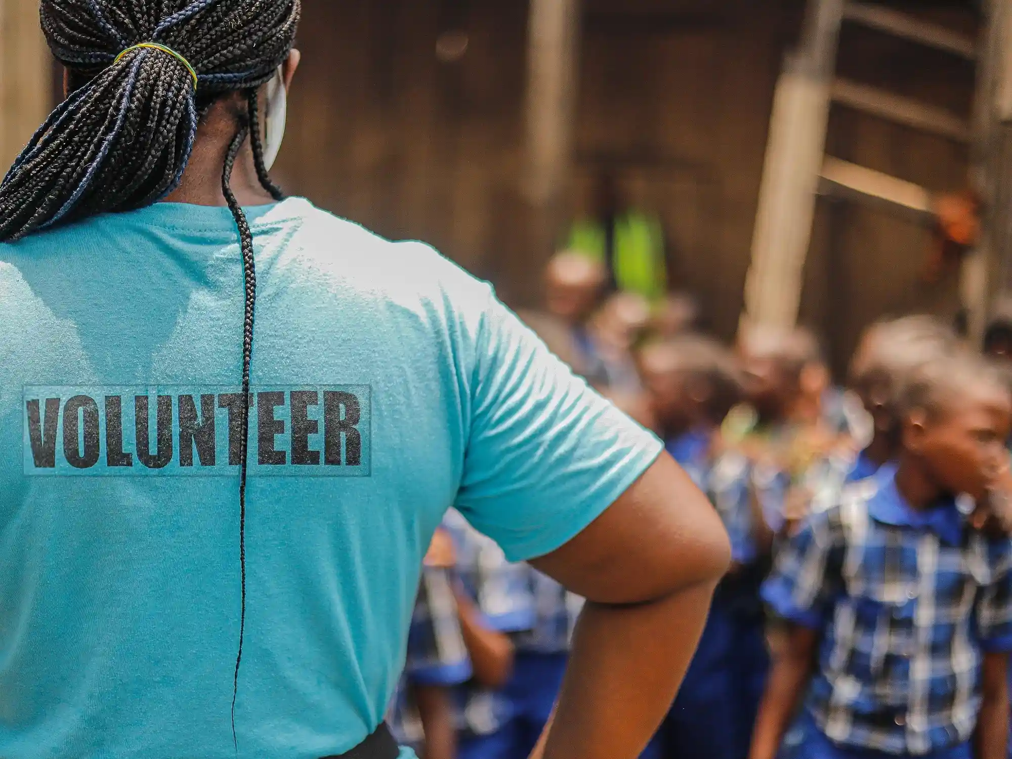 A person wearing a blue "VOLUNTEER" shirt faces a group of children in blue plaid uniforms.