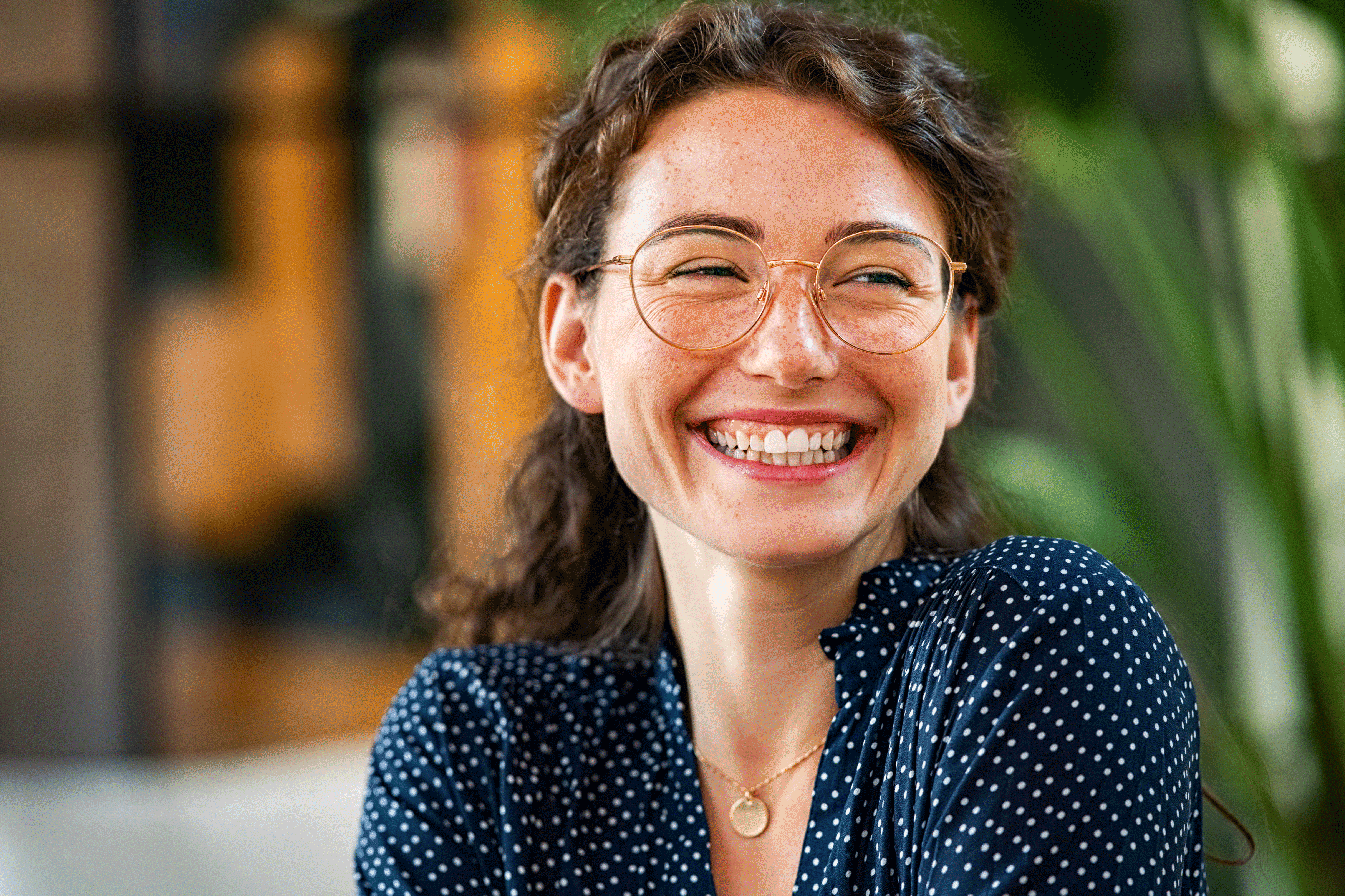 A close-up of a person with wavy brown hair, freckles, and round glasses laughing joyfully. They are wearing a navy blue polka-dot shirt and a gold necklace against a blurred indoor background.