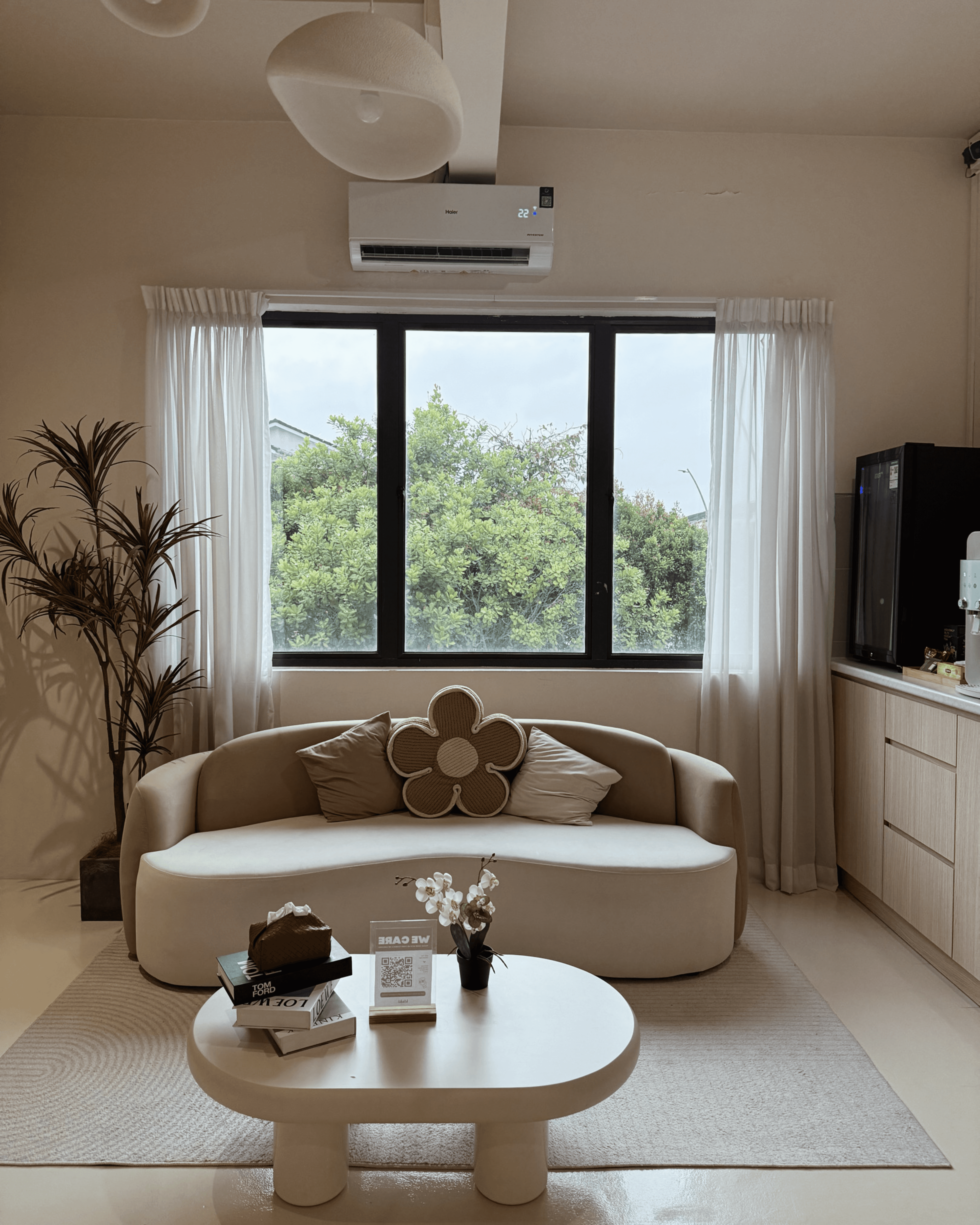 A modern living room featuring a light-colored, curved sofa with a floral accent pillow, a round coffee table with a small plant and decorative books, large windows with sheer curtains revealing greenery outside, and a wall-mounted air conditioner.