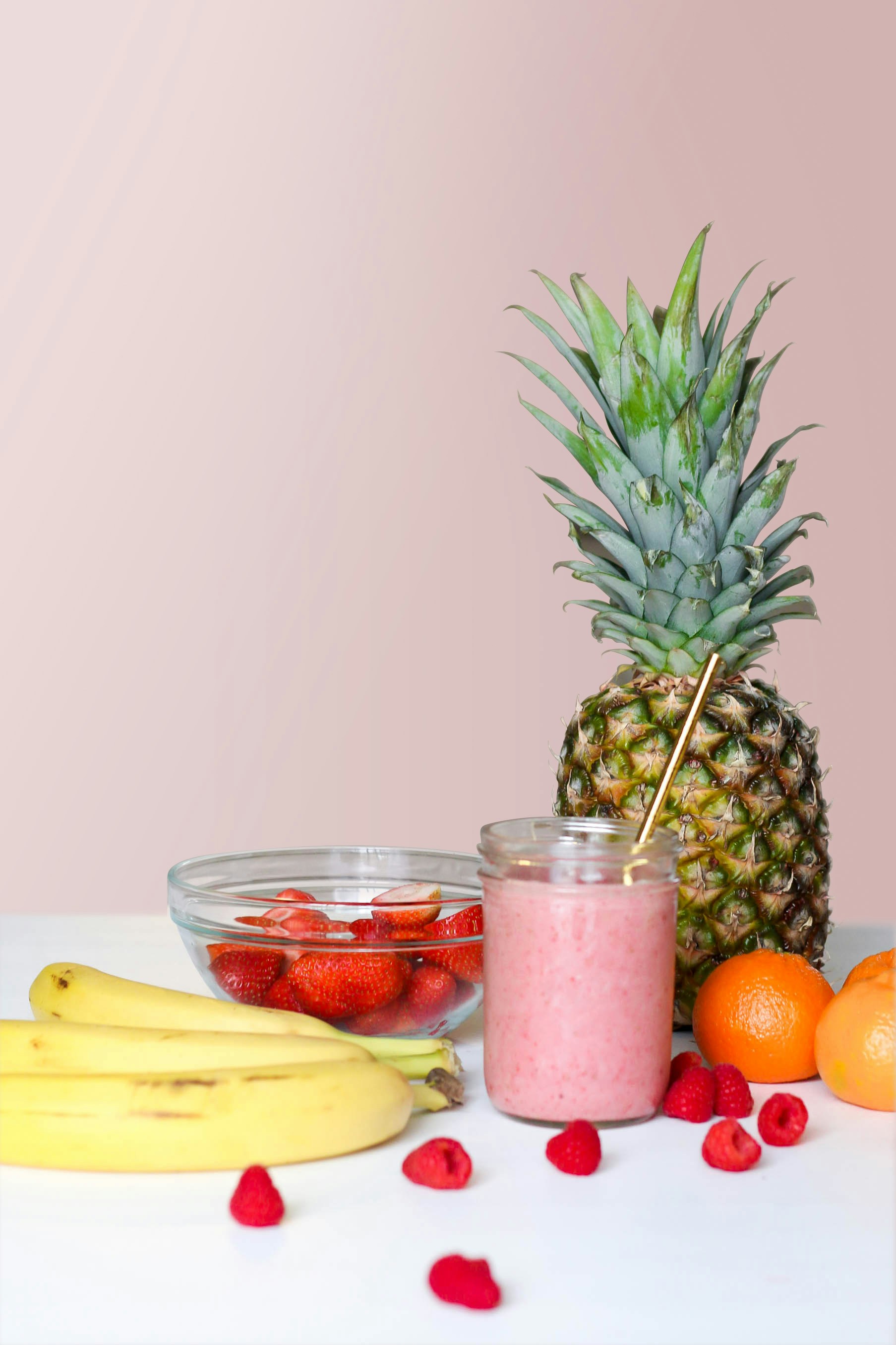 Close up of a smoothie on a table surrounded by fresh fruits banana, pineapple, tangerines, raspberry, strawberry