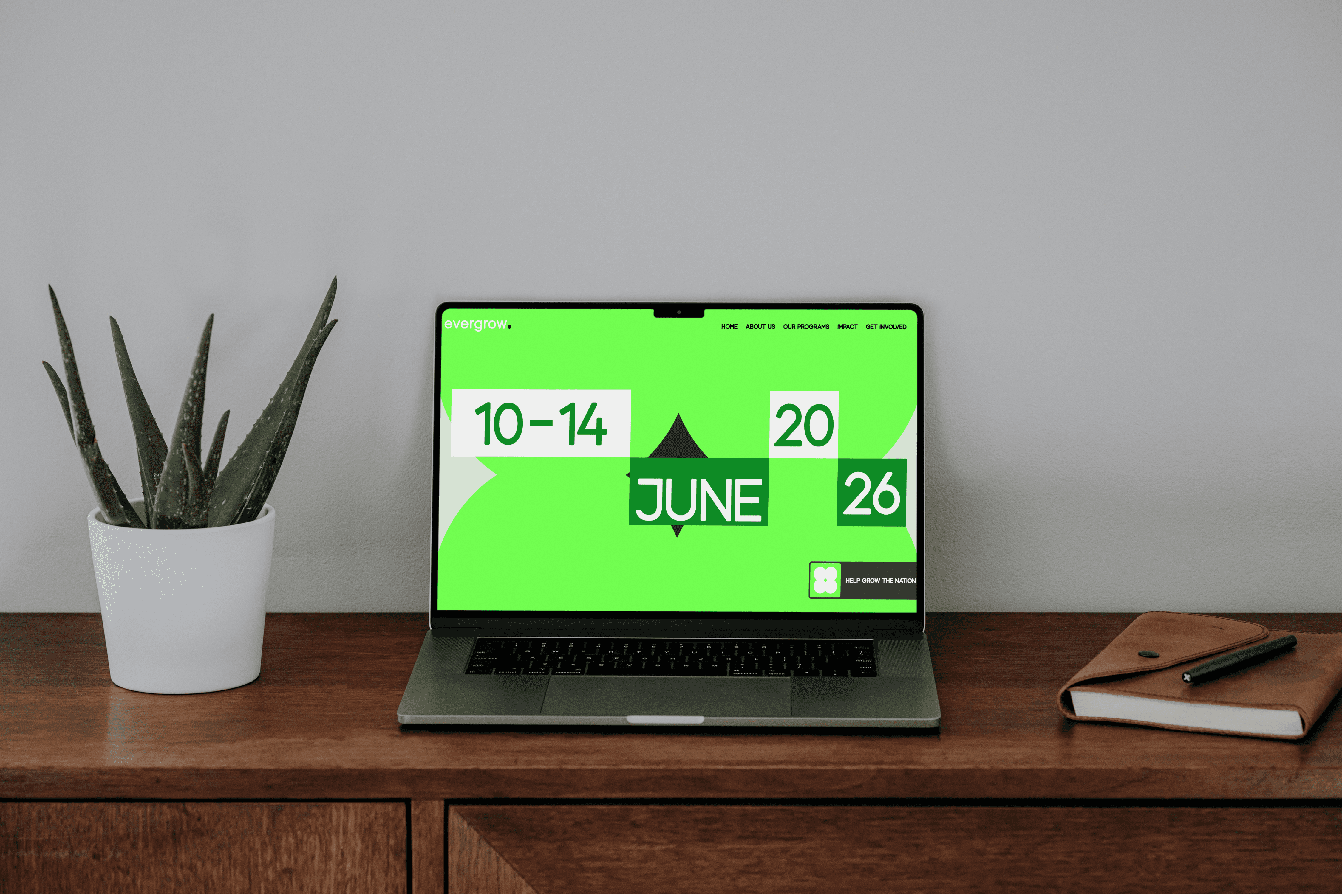 A laptop displaying a vibrant green screen with calendar dates "10-14" and "20-26 June" is placed on a wooden desk next to a potted aloe vera plant and a closed notebook.