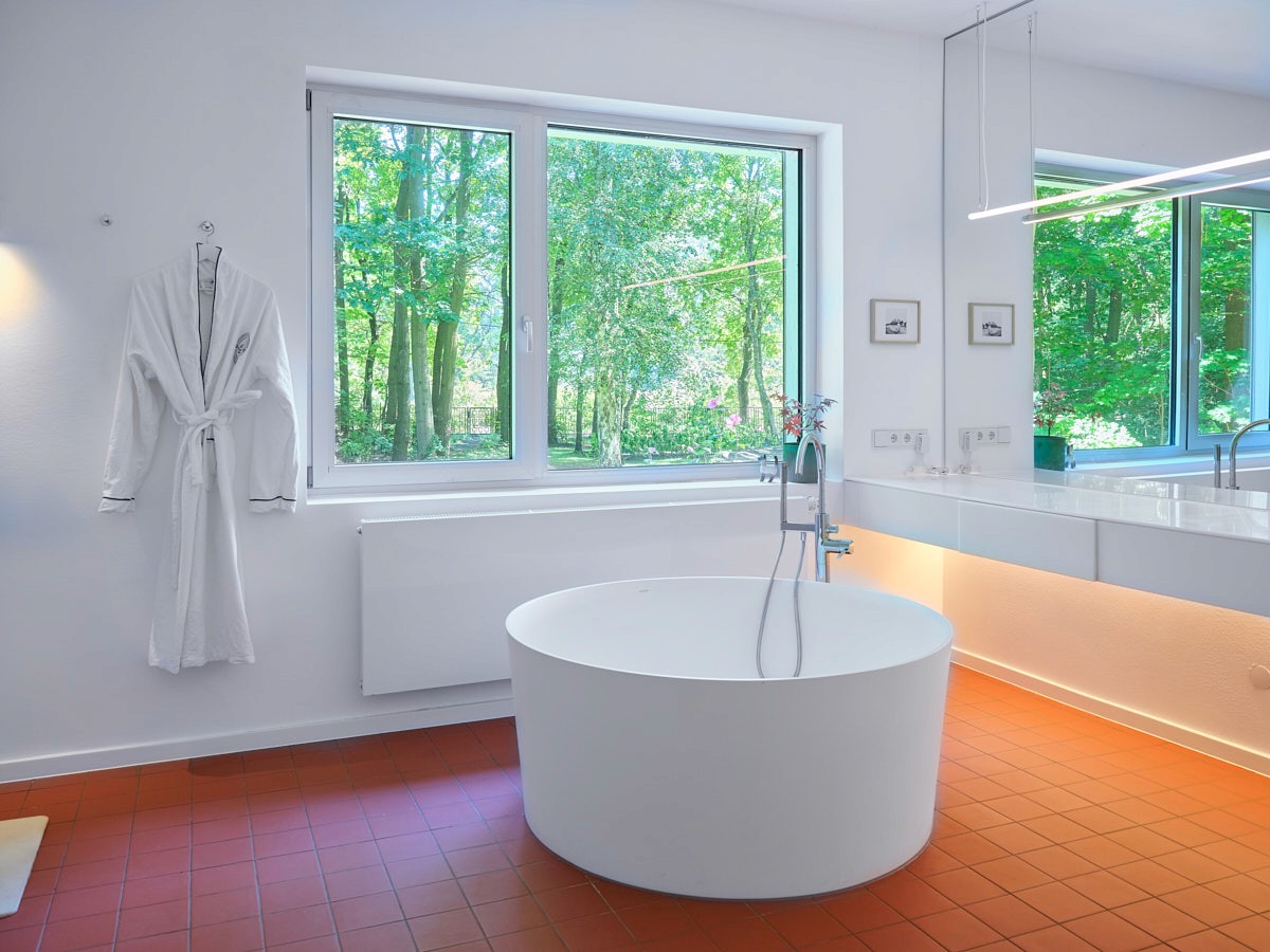 Modernes, helles Badezimmer mit runder freistehender Badewanne, großem Fenster mit Blick ins Grüne und weißem Bademantel an der Wand.