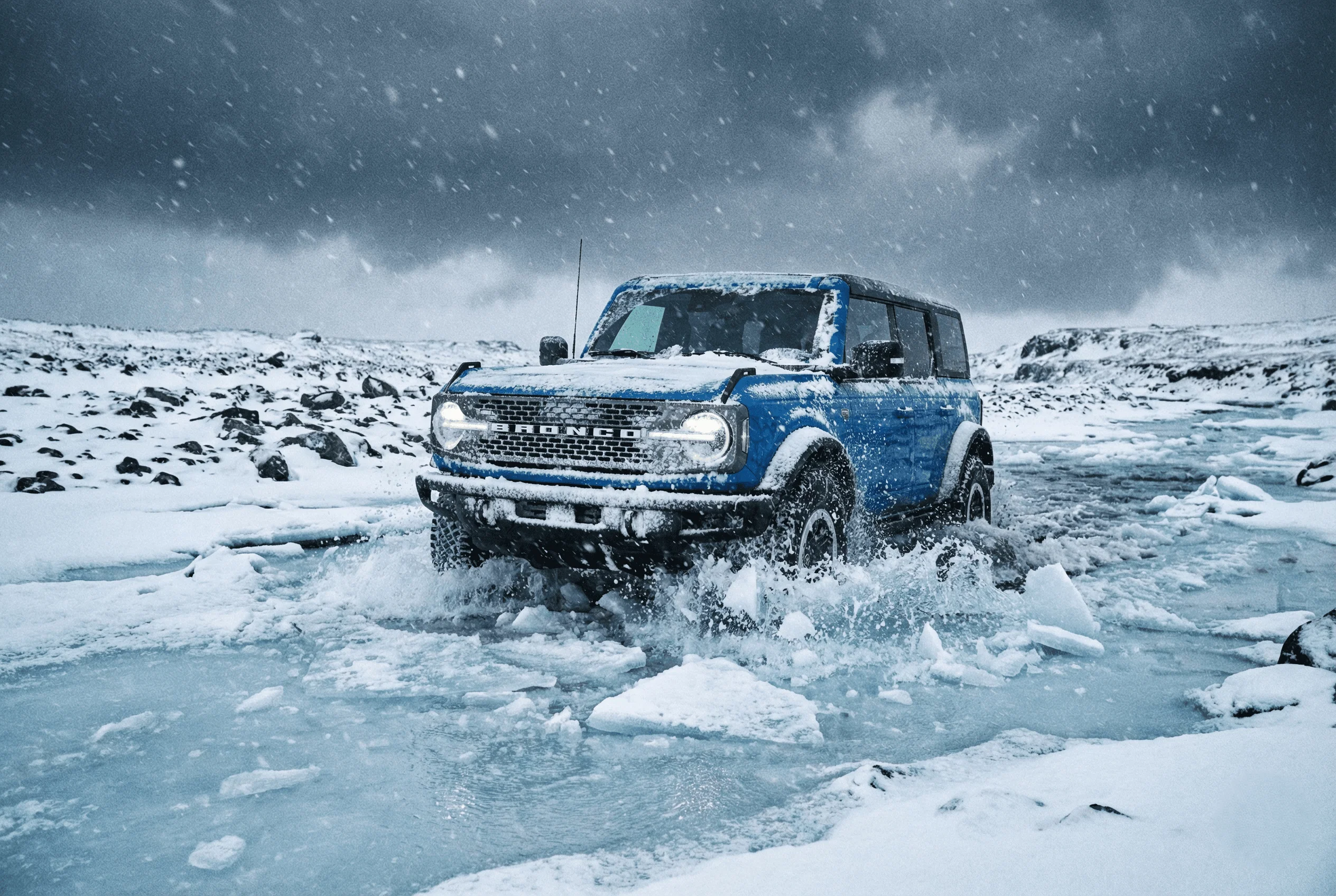 Blue Ford Bronco wading through a frozen river with snowy mountains in the distance