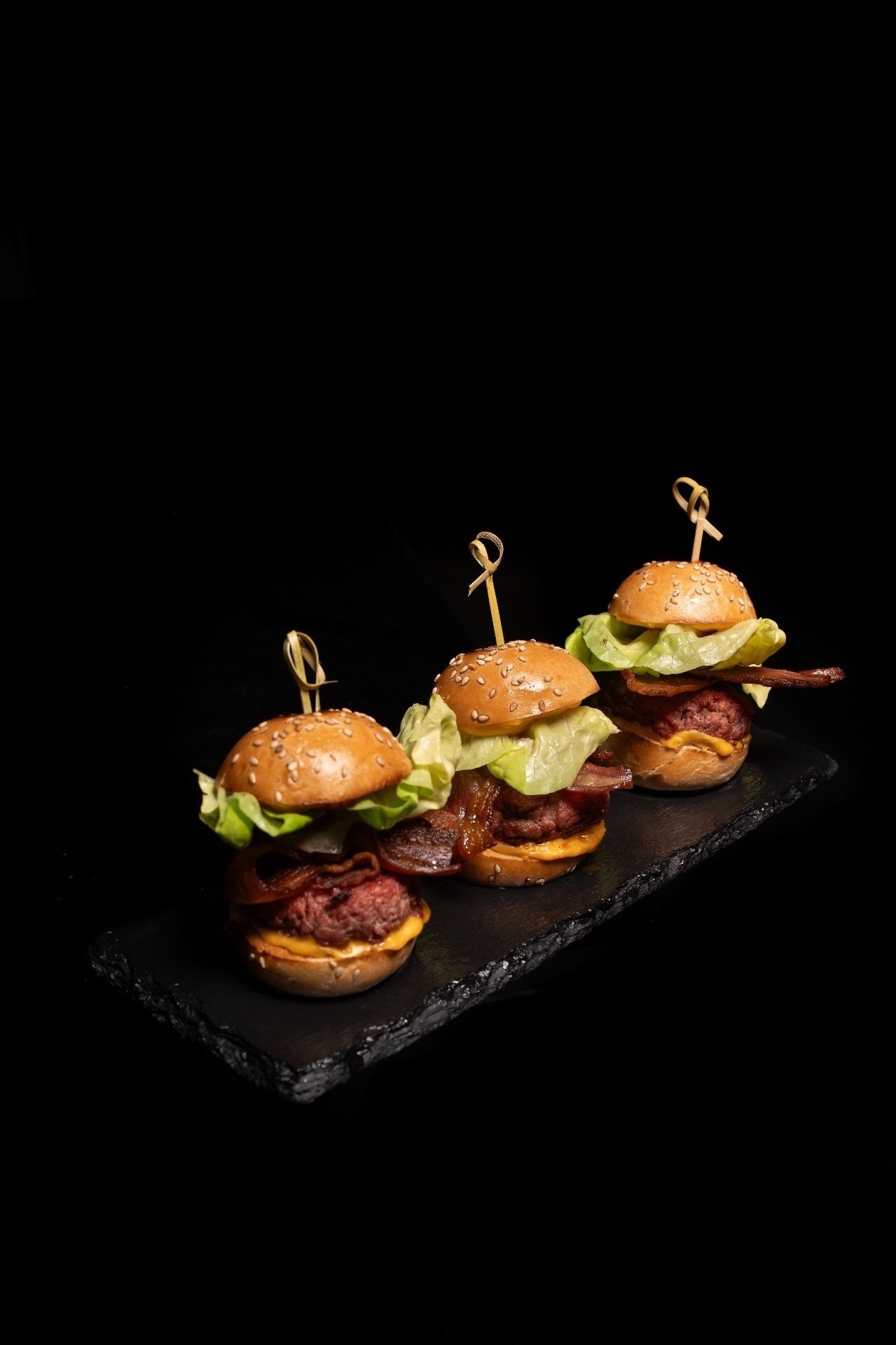 Tre mini burger di manzo su vassoio in ardesia scura, con sfondo nero