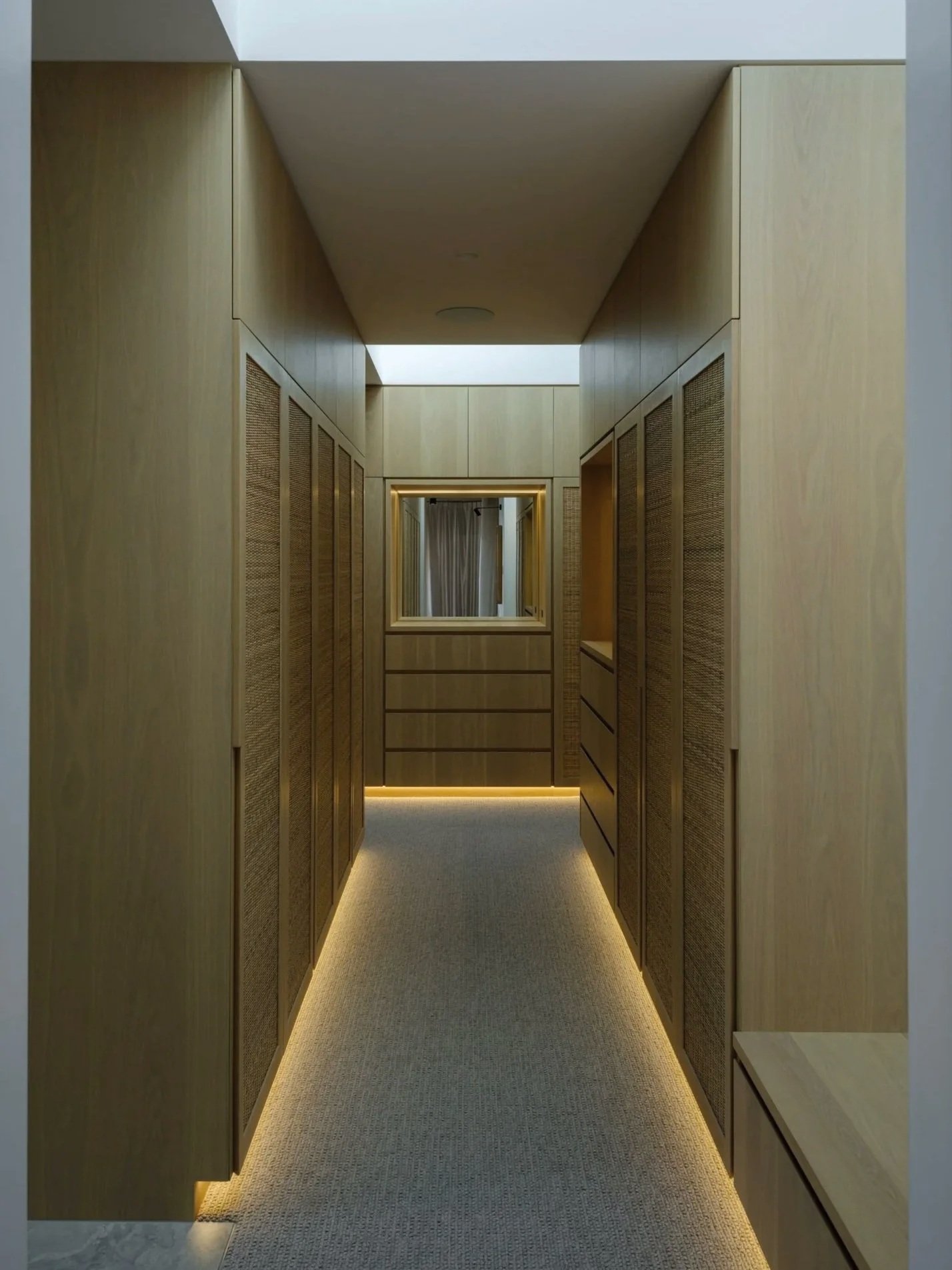 Walk-in robe with full-height timber cabinetry, soft underlighting, and central mirror at the end of a narrow passage.