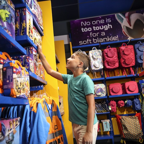 A boy in a green shirt reaches for a toy on a store shelf filled with colorful toys and backpacks. A sign reads, "No one is too tough for a soft blankie!"