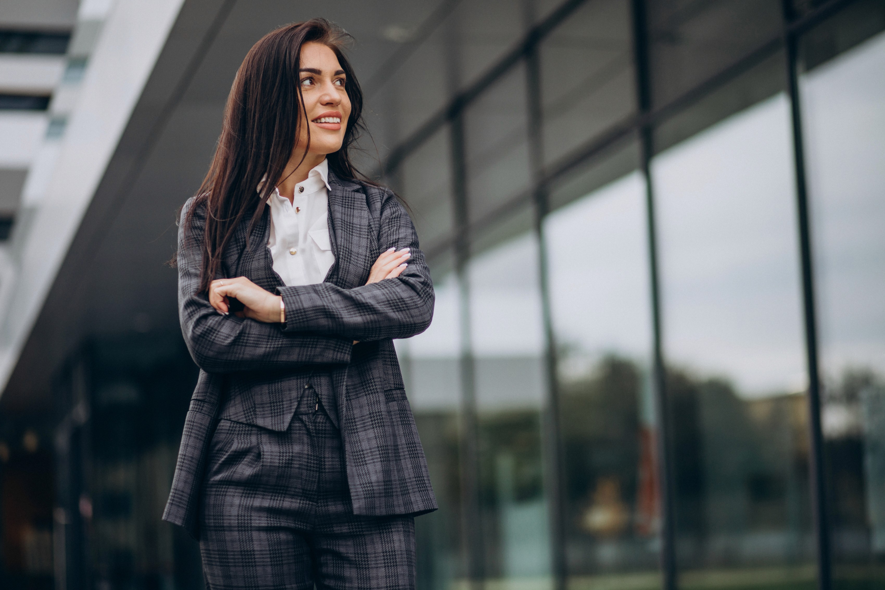Woman in a gray plaid suit stands confidently with arms crossed outside a modern glass building, reflecting professionalism and success.