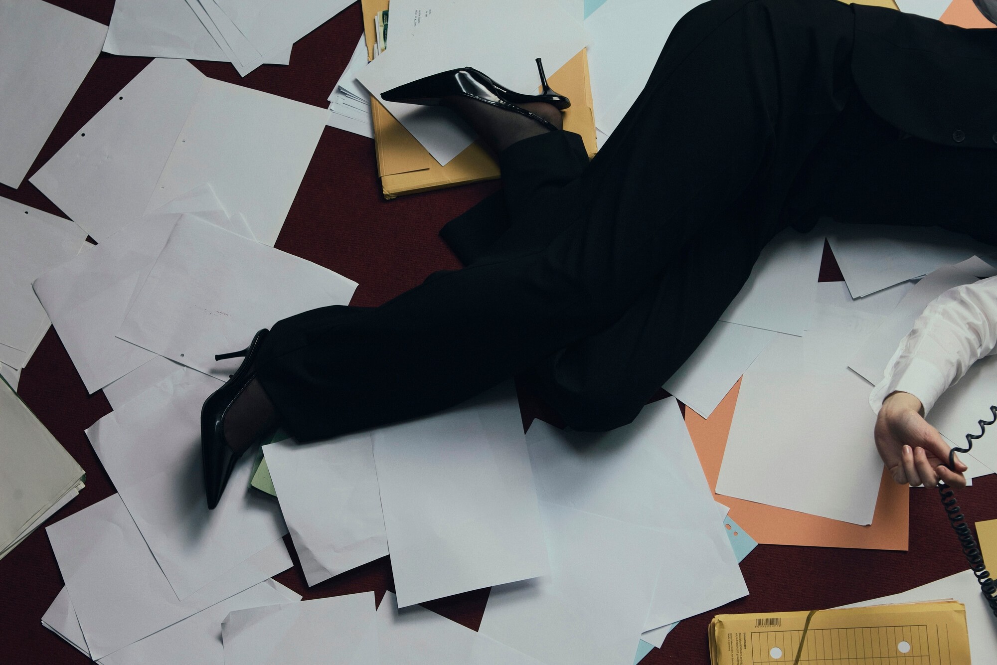 A person in business attire lies amidst scattered papers and folders on a red carpet, holding a phone receiver. The scene conveys chaos and stress.