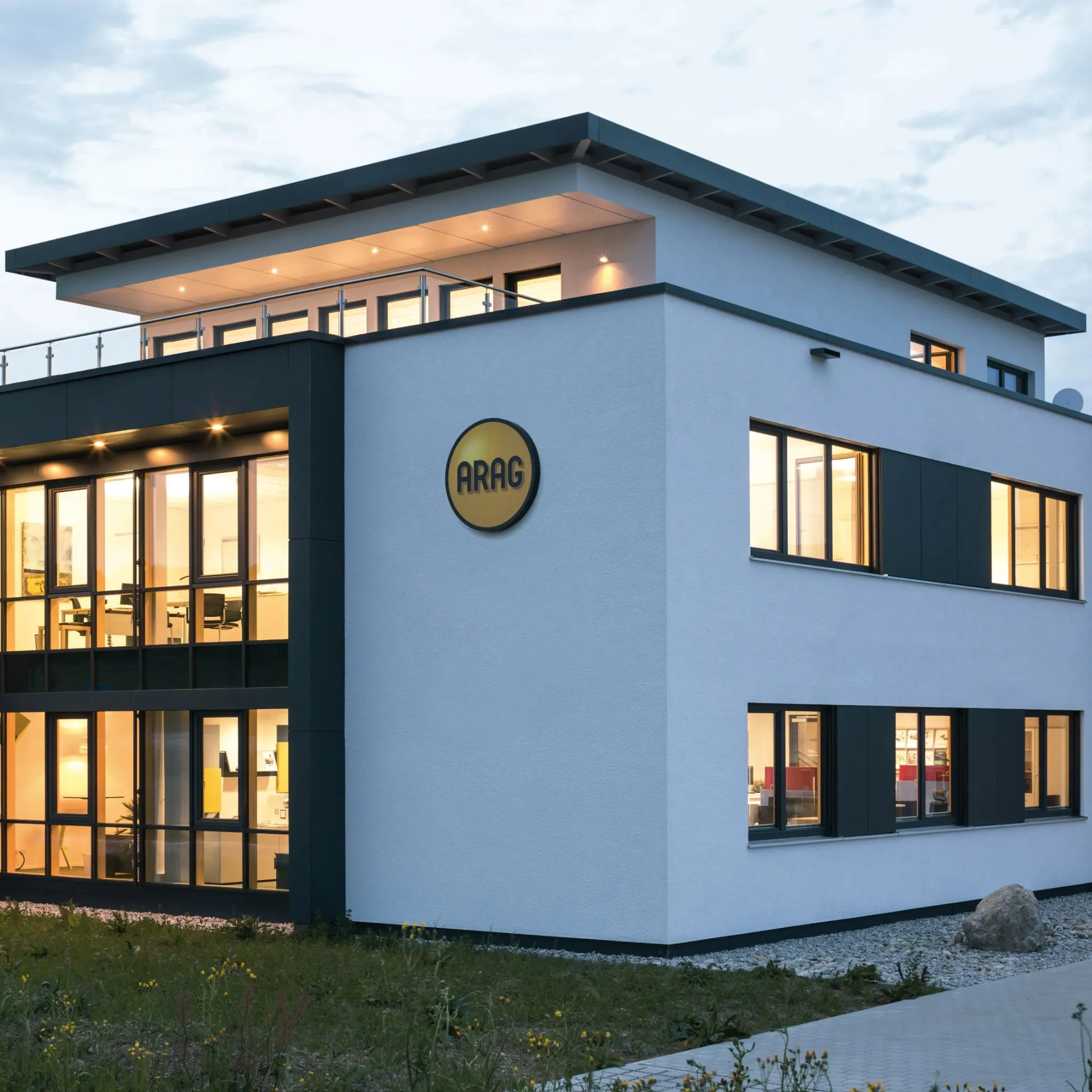 Modernes Bürogebäude mit großflächigen Fenstern und dem ARAG-Logo an der Fassade, in der Dämmerung beleuchtet.