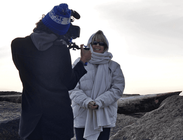 A person in a blue knit hat with "FIRE 2300" printed on it and a black jacket films a person wearing a white hooded jacket and sunglasses. The person in the white jacket holds a small object in their right hand. They are standing on a rocky surface with a cloudy sky in the background.