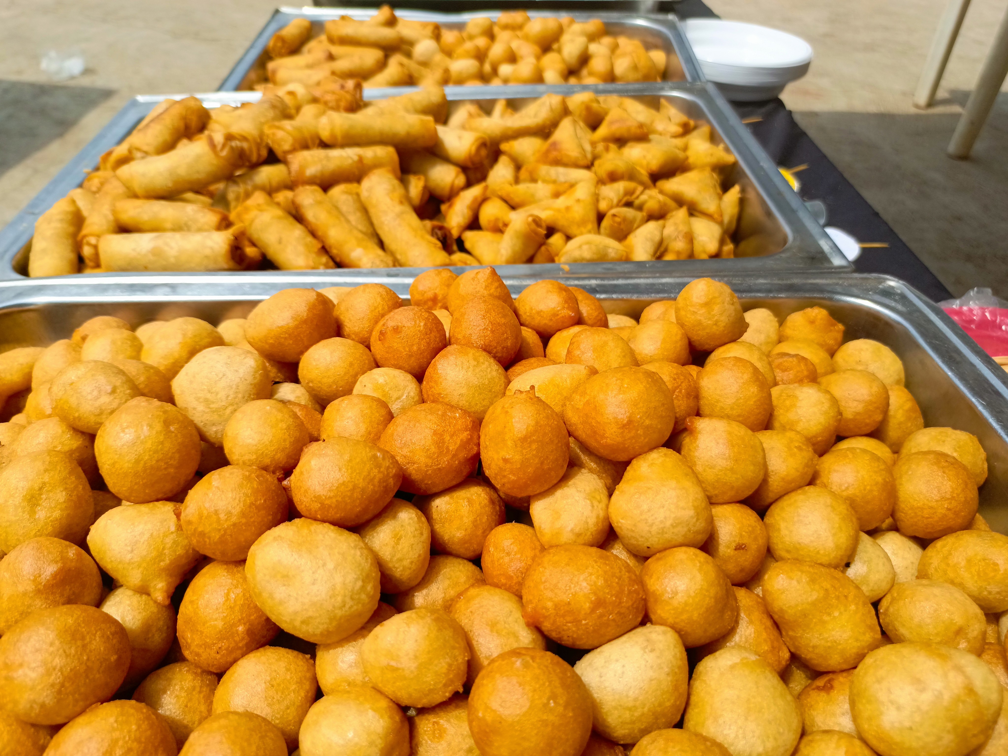 Various fried foods in buffet trays.  Includes spring rolls, samosas, and small round fried balls.