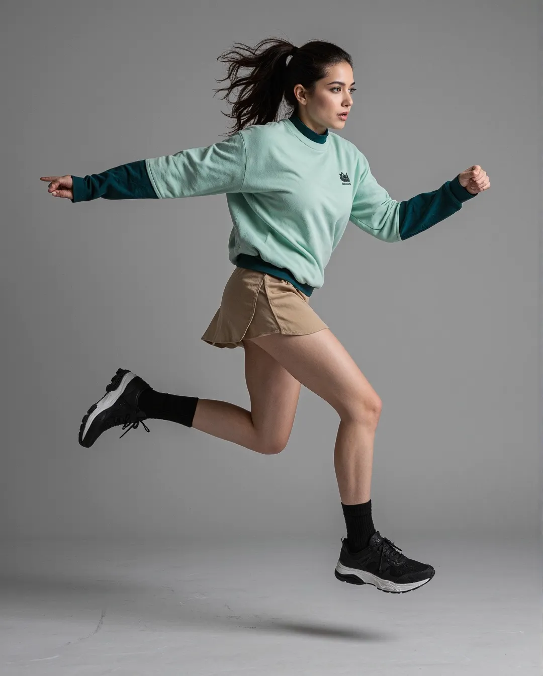 Person in mid-jump wearing mint green sweatshirt with dark sleeves, beige mini skirt and black sneakers against neutral gray background