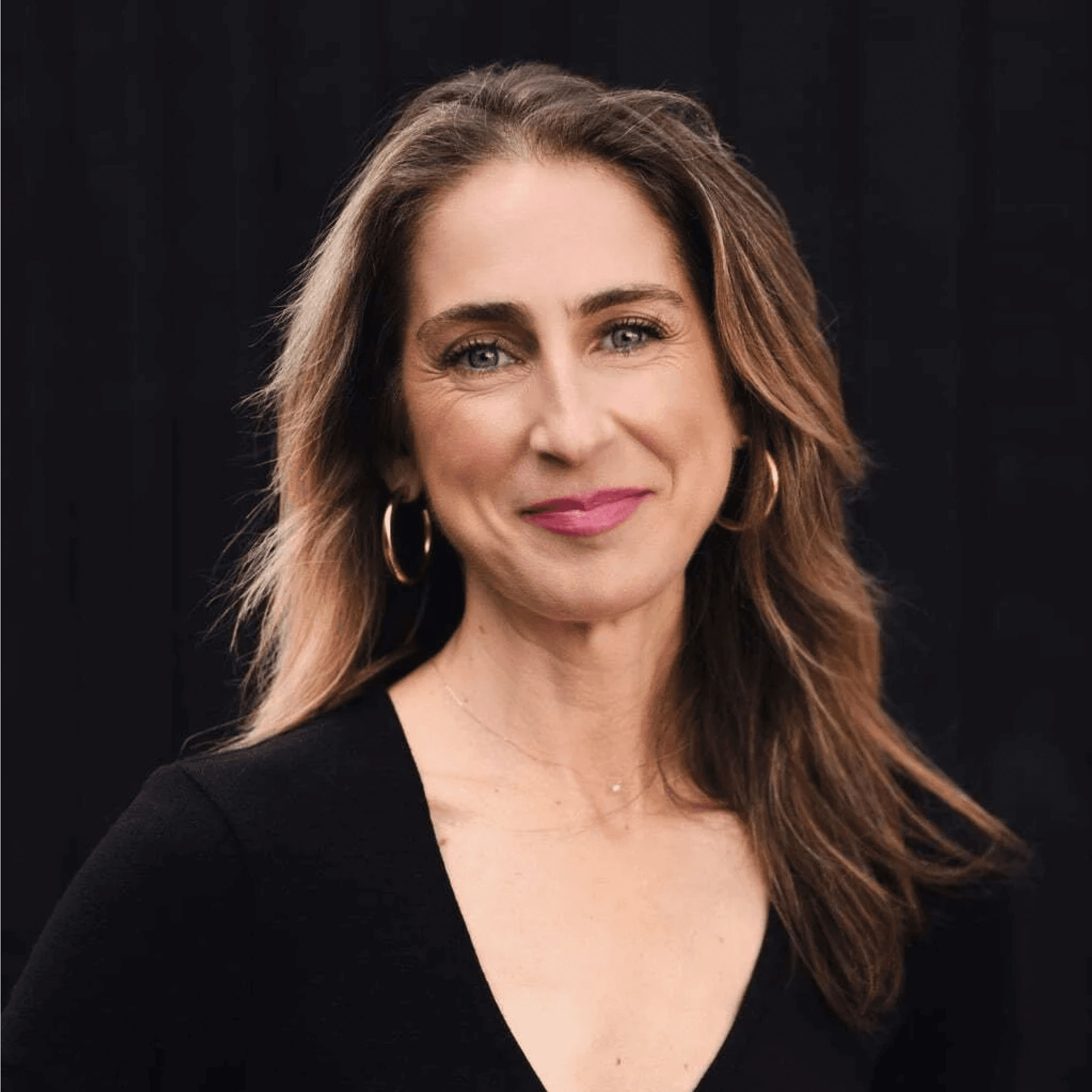 A woman with wavy brown hair smiles softly against a dark background, wearing a black top.