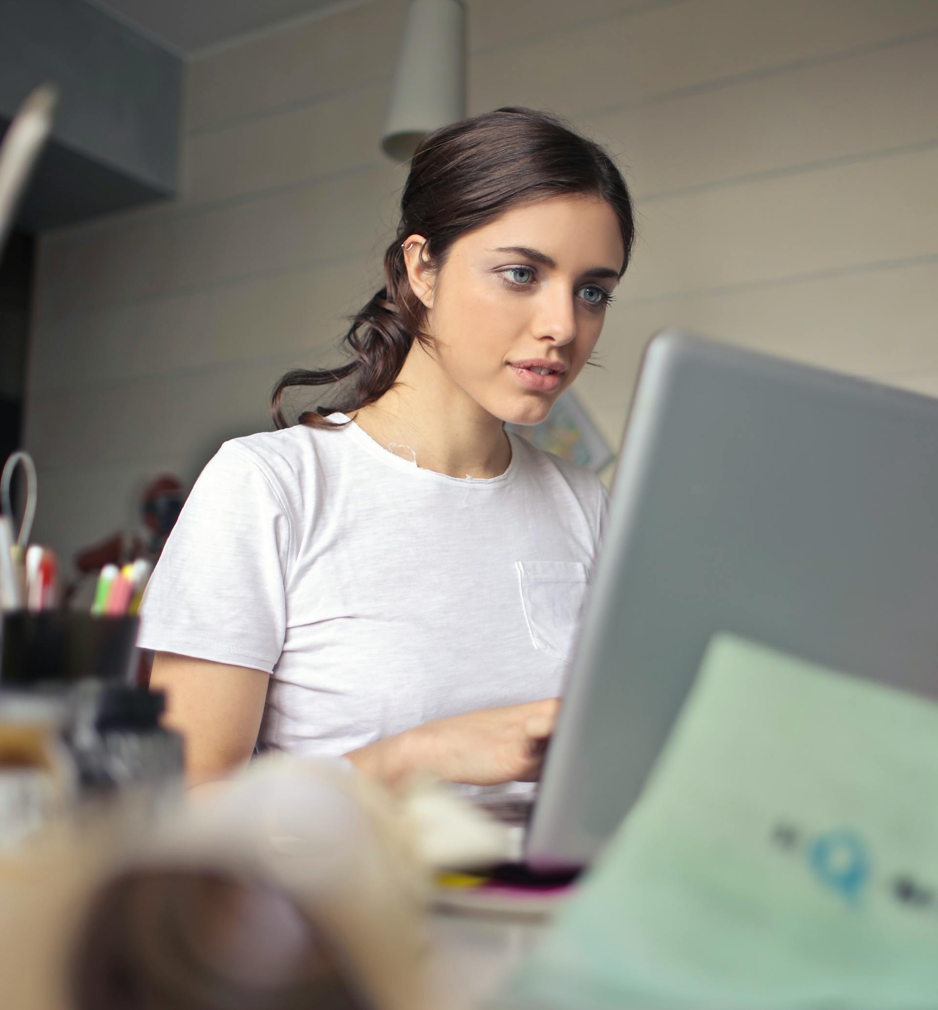 A young woman with dark hair tied back, wearing a white t-shirt, is concentrating while typing on a laptop at a cluttered desk.