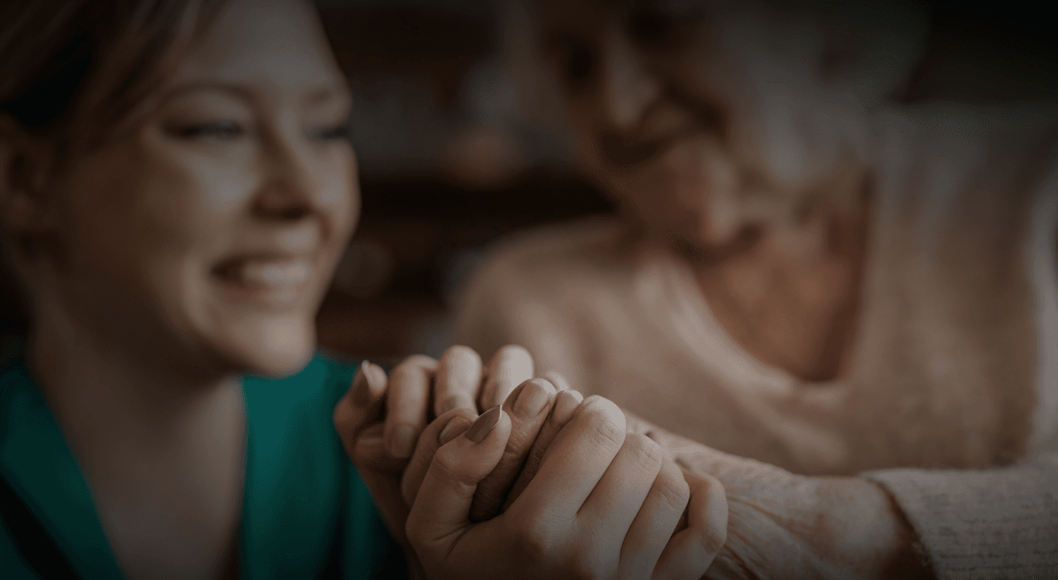 Young female caregiver sits beside older adult, both smile with hands embraced