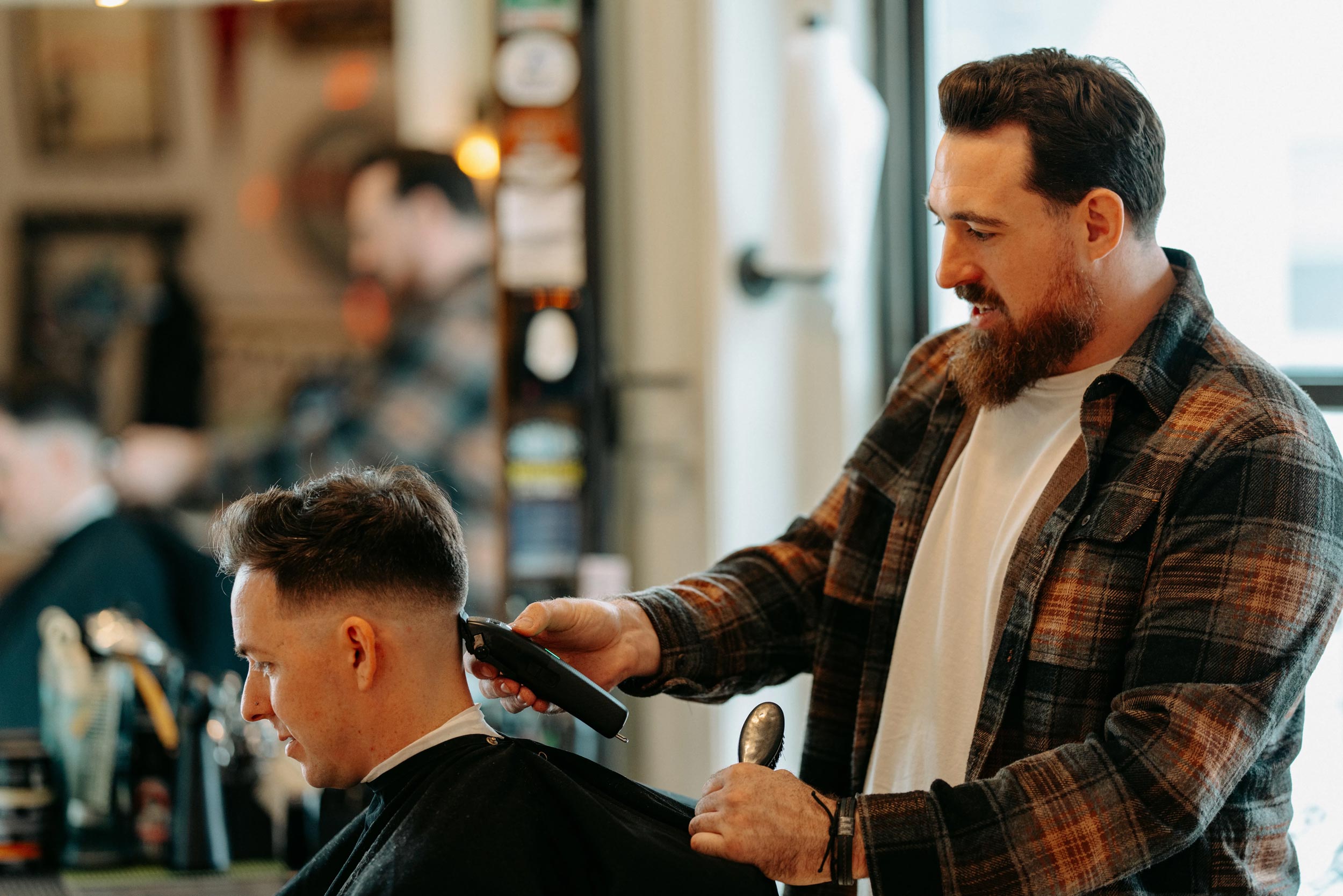 A male barber with a full beard and a plaid shirt is using clippers to cut the hair of a male client, who is seated and draped with a black cape.
