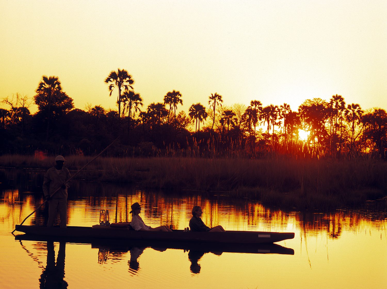 A poler guiding a mokoro through the Okavango at sunset, palms silhouetted beyond
