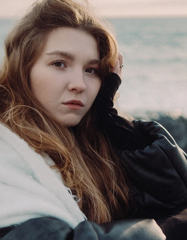 A young woman with long brown hair gazes thoughtfully into the distance. She's wrapped in a cozy black and white coat by the seaside, under soft, natural light.