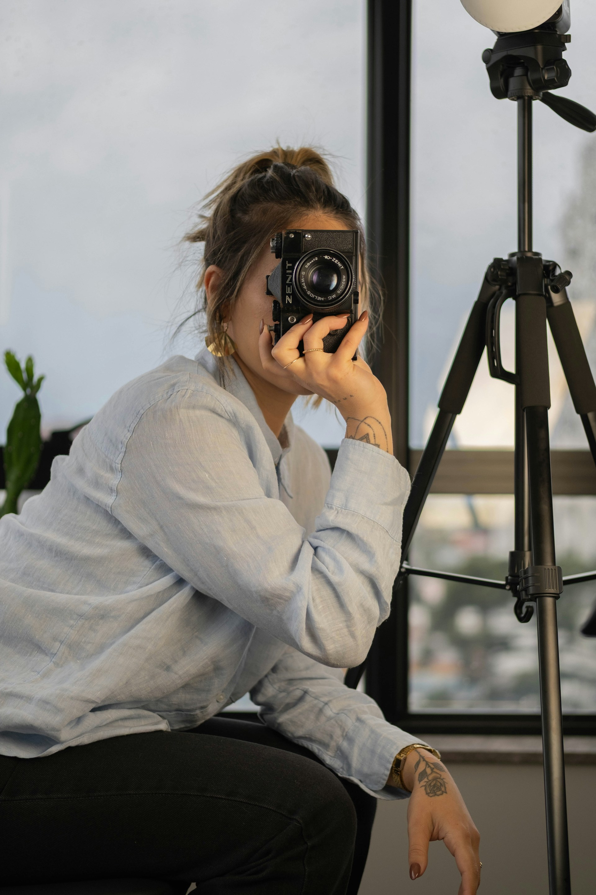 A woman with her hair pulled back is holding a black vintage-style camera up to her eye, preparing to take a photo, with a black tripod visible to her right.
