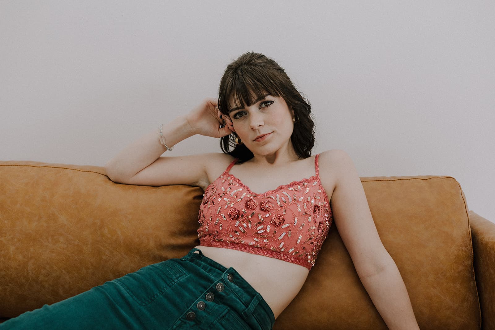 A model reclining on a tan couch in a pink embellished top and green pants, captured in the natural light at Revelator Studio, a photography studio in Shreveport.