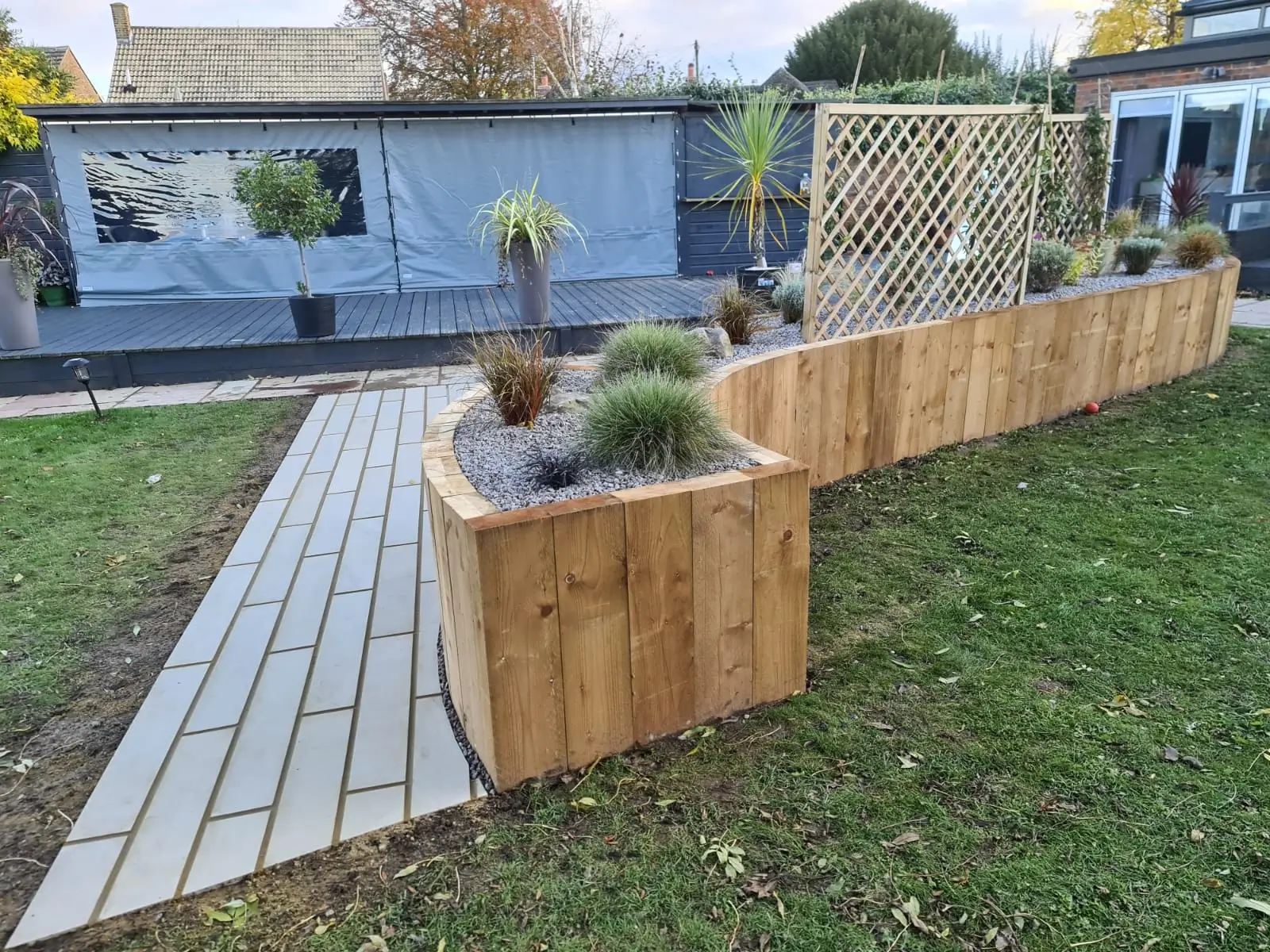 Garden with wooden raised planter beds and a stone pathway, surrounded by grass and a blue building in the background.