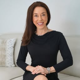 A woman with long dark hair sits on a sofa, smiling and wearing a black dress in a bright, airy room.