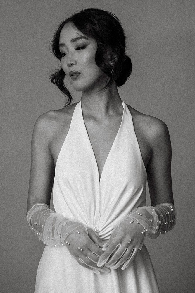Black-and-white bridal editorial portrait of a woman in a plunging halter-neck gown with sheer pearl-embellished gloves, hands clasped, fine art photography by Eva Sitko