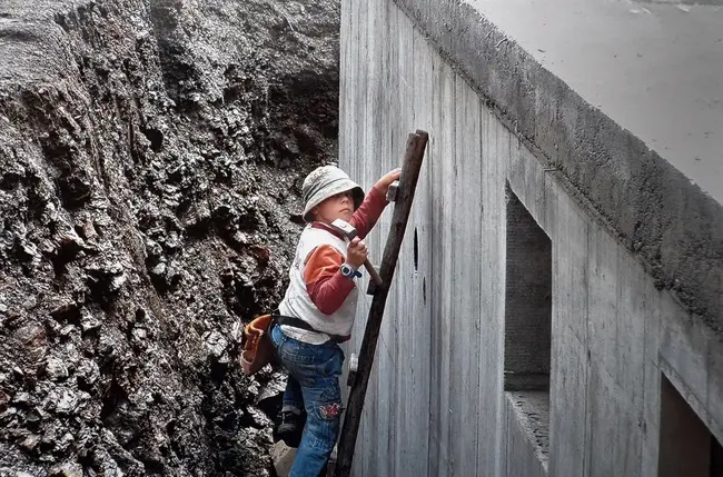 Einer der Schmoltner-Söhne als Kind mit Hammer und Werkzeuggürtel auf einer Holzleiter an der Betonwand des Rohbaus.