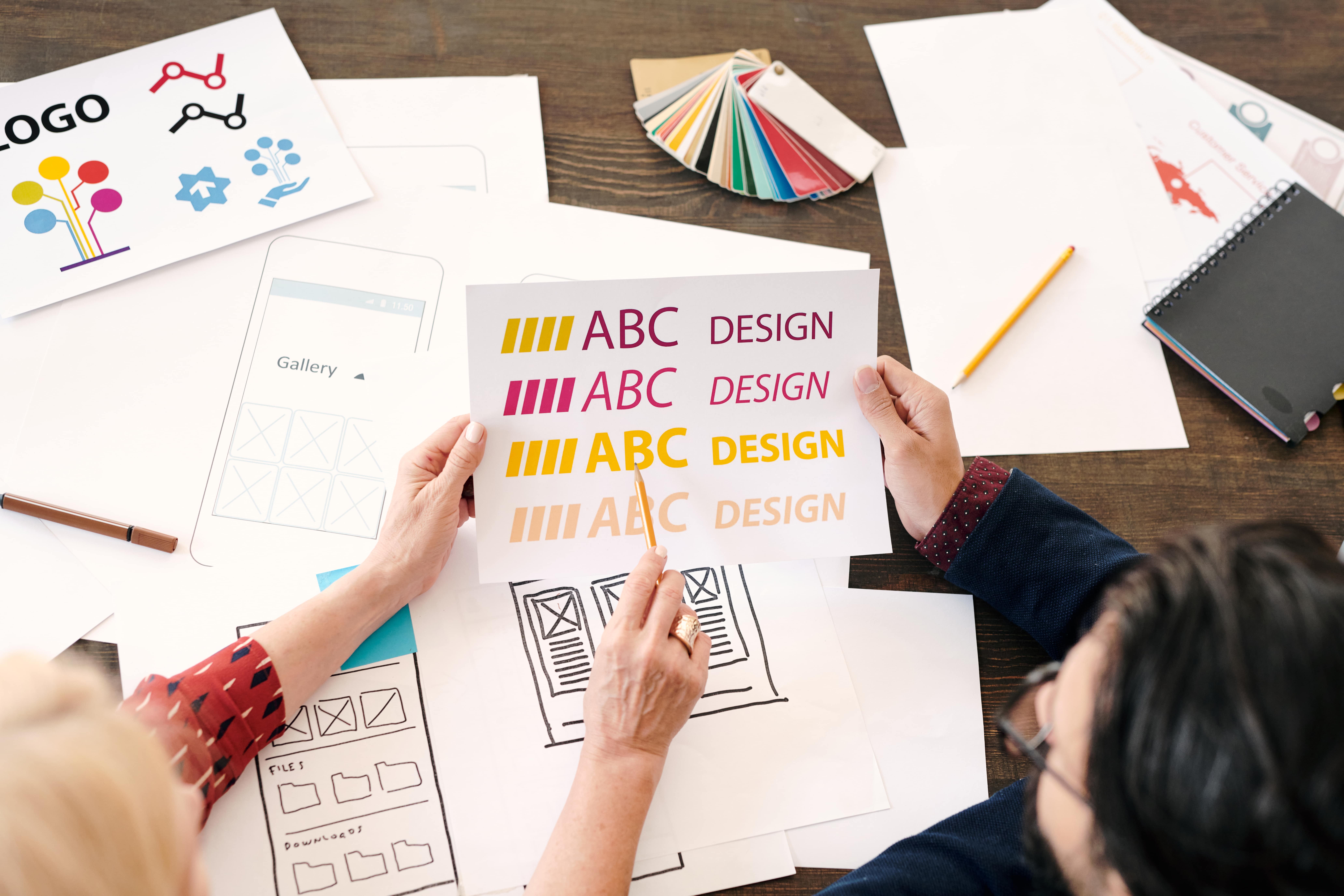 Two people review color and font options for a design project at a table with sketches, color swatches, and logo concepts spread out.