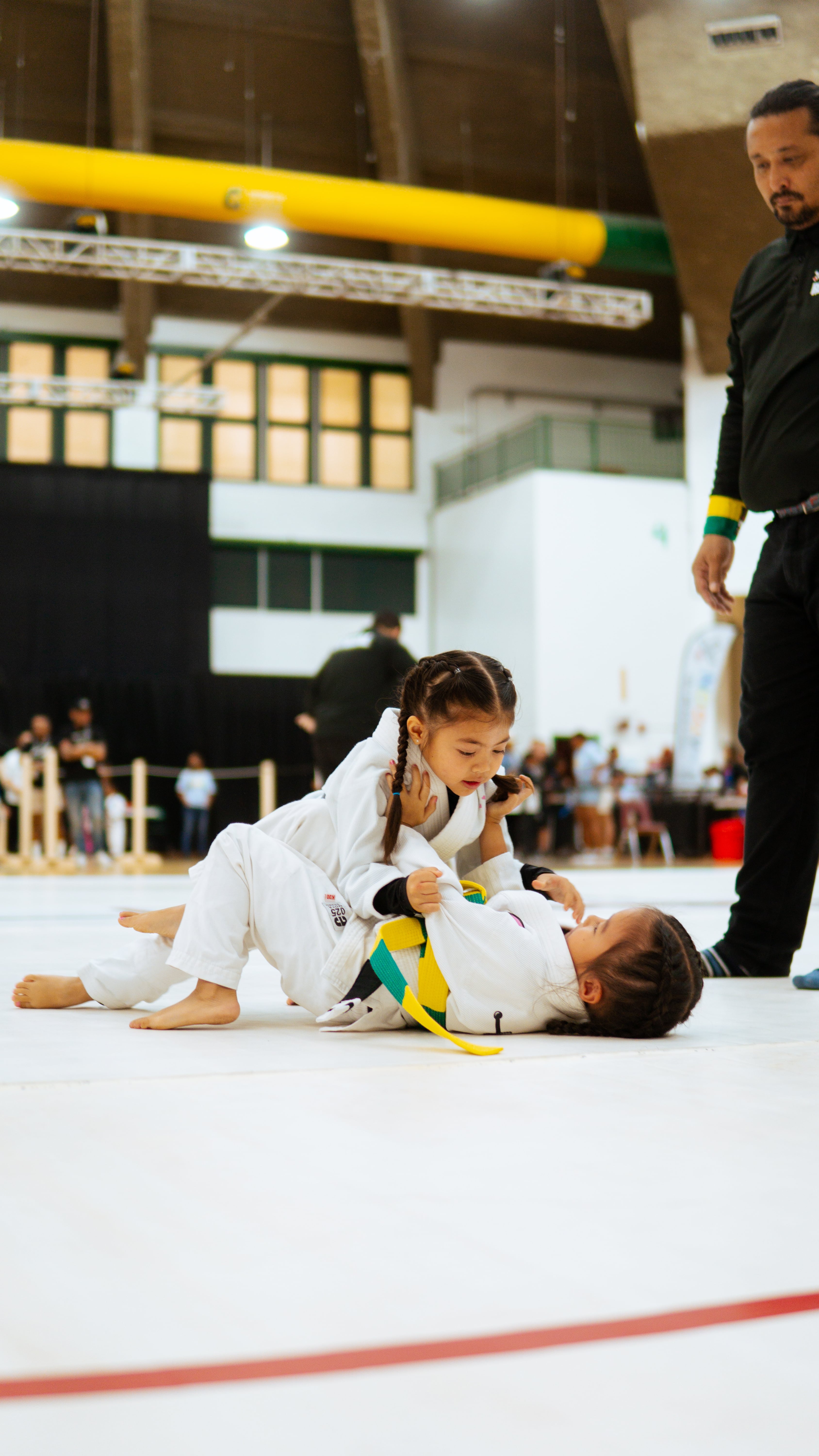 Two young children in white jiu-jitsu uniforms compete on the mat, one performing a hold on the other during a Pacific Grappling Events tournament.