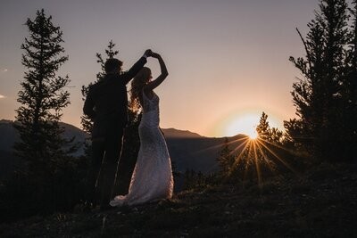 The couple dance in silhouette as the sun sets between the evergreen trees.