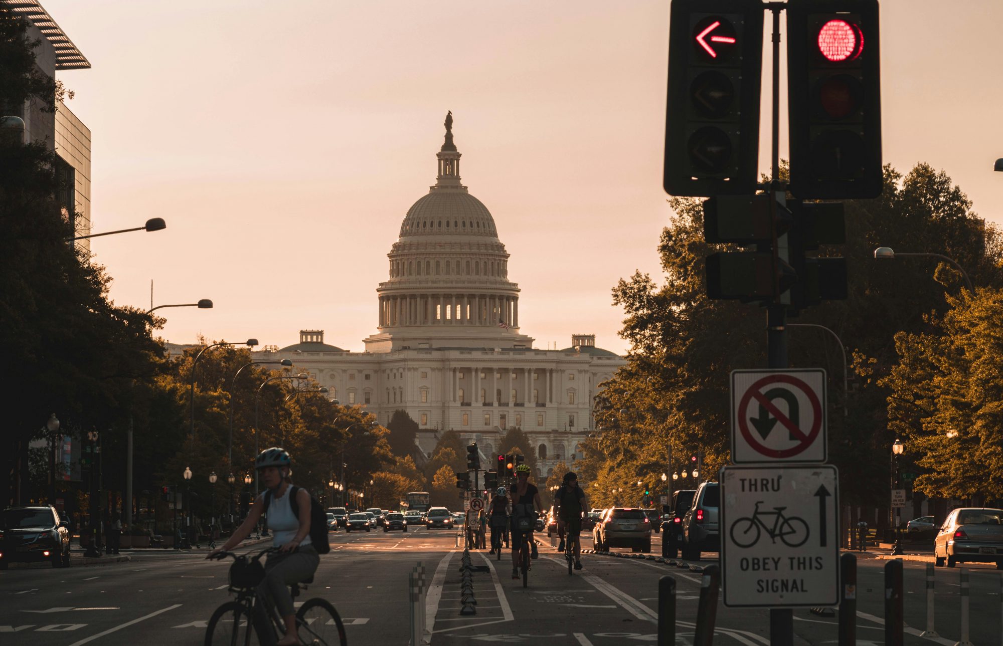 Silhouetted pedestrians cross a street in front of a large dome building during sunset, with traffic lights visible.